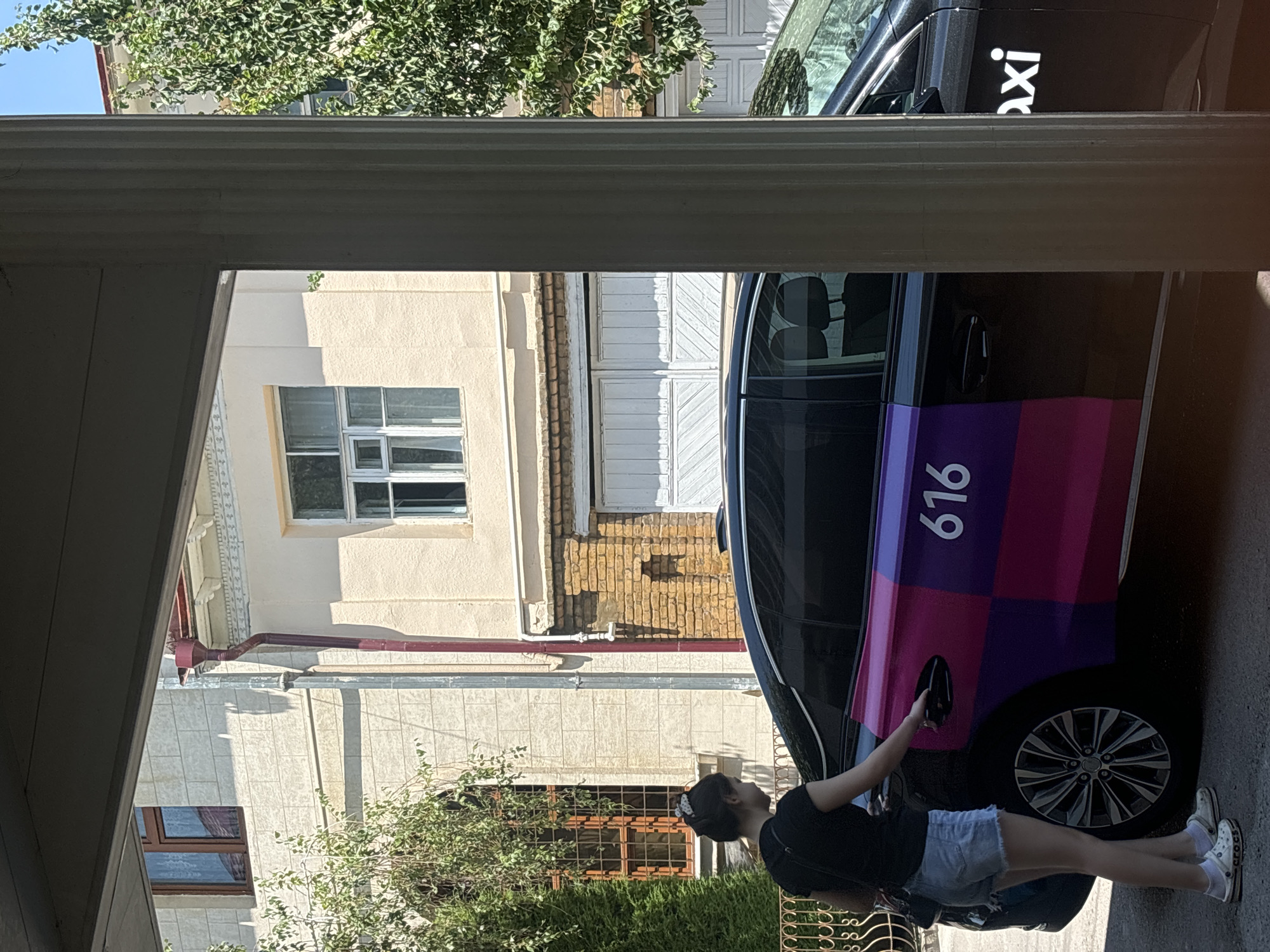 A young woman in a black t-shirt and denim shorts is opening the rear passenger door of a black taxi. The taxi has a distinctive purple and pink checkered pattern on its rear quarter panel with the number "616" in white lettering. The word "axi" in white lettering is visible on the rear door. The scene appears to be in a residential area, with a multi-story building in the background. The building features light-colored facade with decorative elements, brown window frames, and a red downspout. There are trees and shrubs surrounding the building. The overall impression is a sunny day in an urban setting.