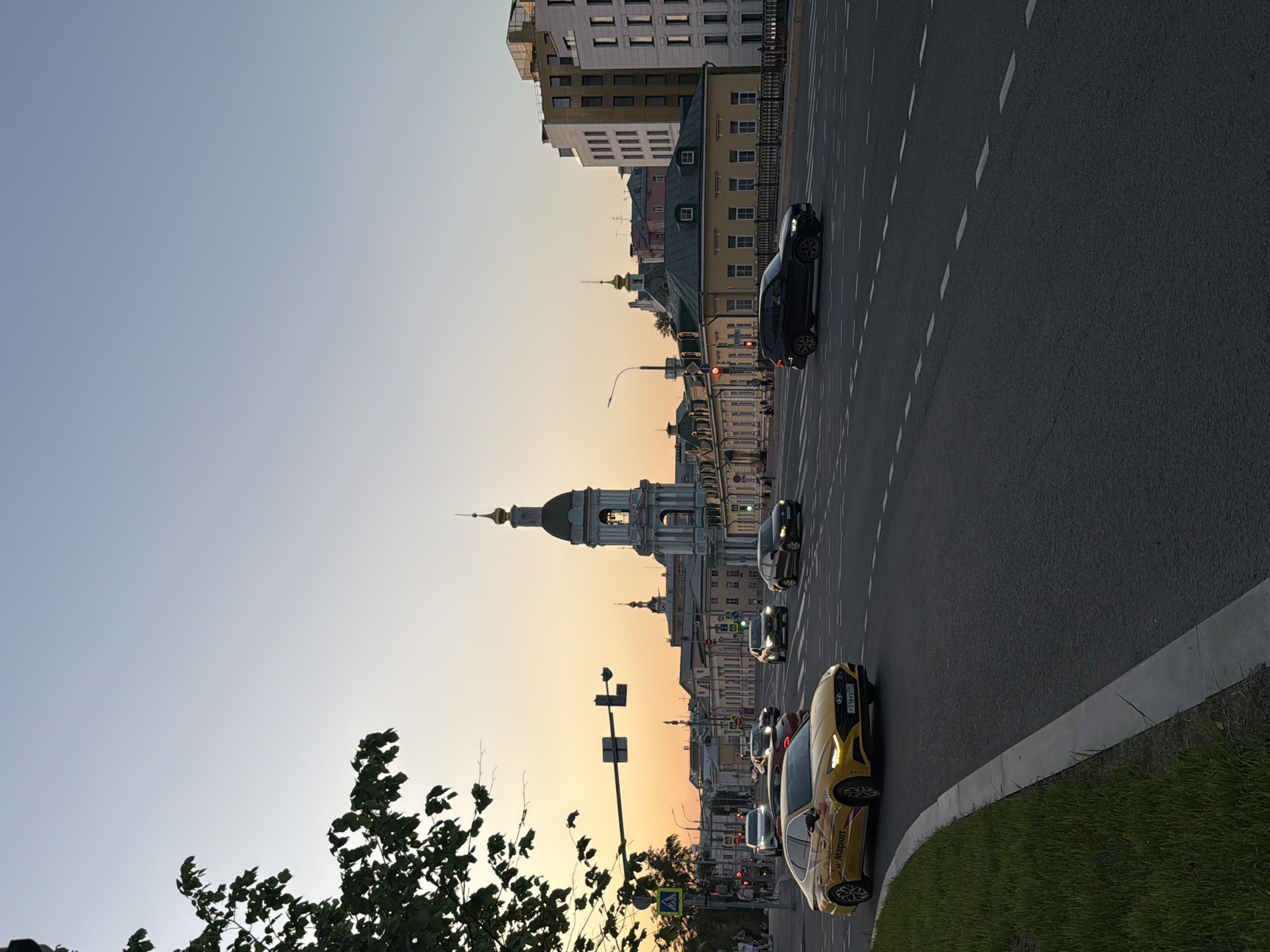 Here is a description of the image:
The image shows a street scene in Moscow, Russia at sunset. Cars are traveling on a multi-lane road. A tall building with a distinctive dome is visible in the background along with several other buildings. Trees and greenery are visible in the foreground and on the road. The sky is clear and the light is soft due to the setting sun.