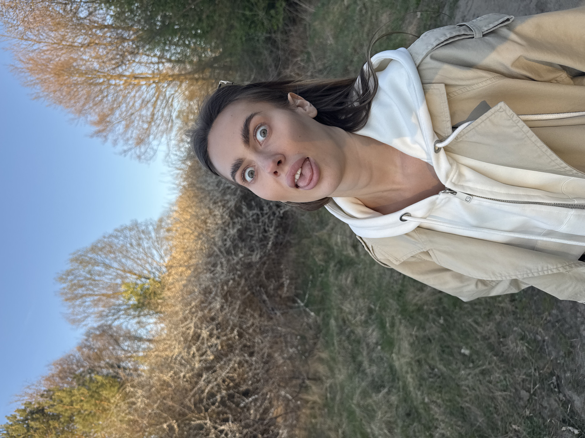 A woman is outdoors, with a facial expression of surprise and her tongue sticking out. She is wearing a white hoodie with a beige jacket over it. The background shows trees with budding leaves against a blue sky. The ground appears to be covered in grass and some dirt patches. The location is uncertain, however, the city of Arciechów is in Poland.