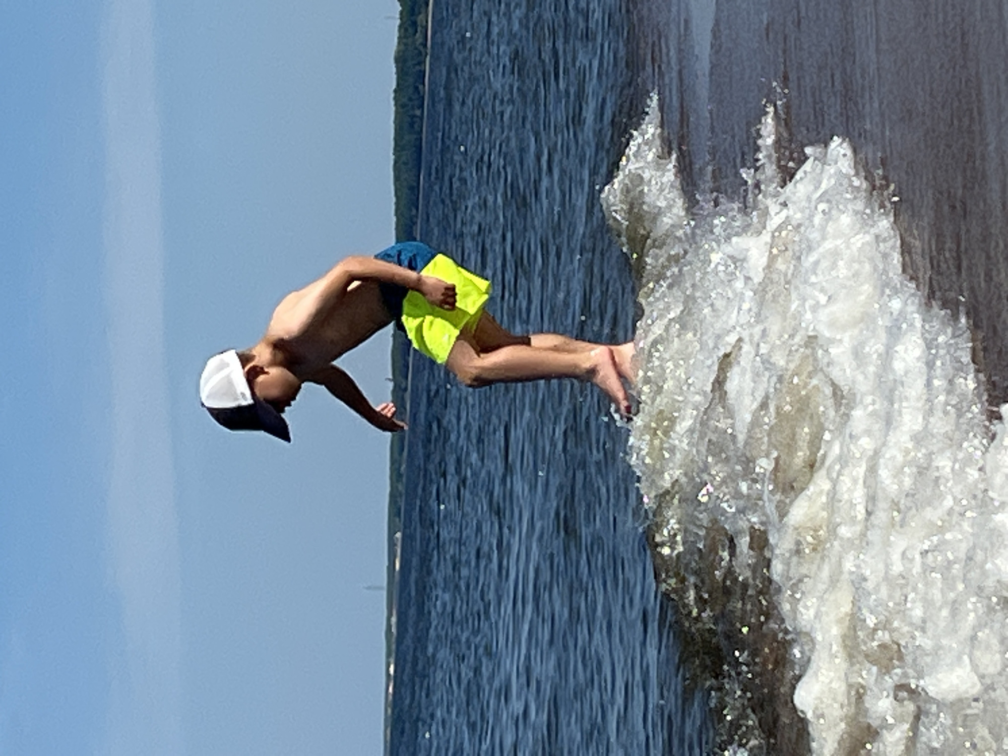 An athletic young boy with a tan complexion and dark hair is standing on a sandy shore at the edge of a body of water. He is wearing a white and dark baseball cap, and lime green swim trunks with a dark blue waistband. The boy is captured mid-action, leaning forward with his right arm extended downwards and his left arm bent, as if he is balancing or about to jump. He is barefoot and appears to be standing on a small wave that is breaking onto the shore. The water is a deep blue, and a line of trees is visible in the distance across the water, forming the horizon. The sky is a clear, bright blue. The scene is bathed in bright sunlight, casting subtle shadows and highlighting the texture of the water and sand. The image is taken from a low angle, emphasizing the movement of the waves and the boy's posture.