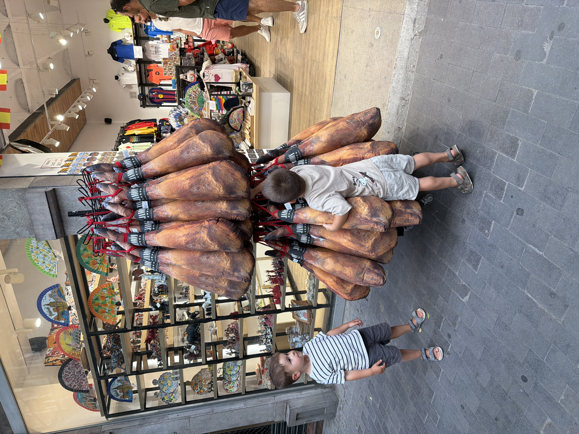 In an outdoor marketplace, two young boys stand on a cobblestone sidewalk in front of a shop. The boy on the left, wearing a striped t-shirt and shorts, looks towards the right. The boy on the right, dressed in a light-colored t-shirt and shorts, is reaching out to touch a large display of cured hams. Behind the hams, the shop's interior is visible, featuring shelves stocked with colorful fans and various trinkets. In the background, two men are browsing the merchandise. The scene suggests a tourist-oriented location, possibly in Spain, given the prominent display of cured hams.