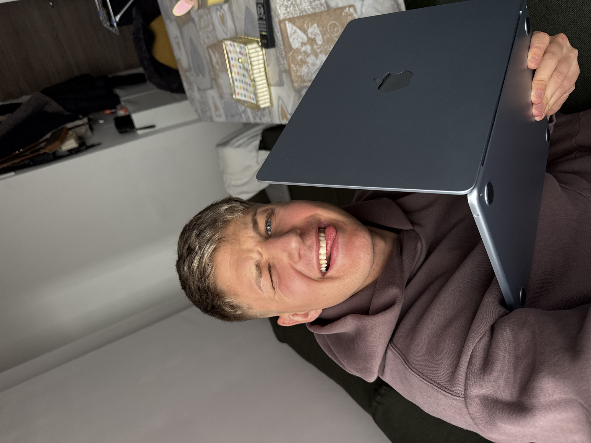 A man is reclining and holding a closed, dark-colored Apple laptop in front of his face. The man is fair-skinned, with short, light brown hair and blue eyes. He is wearing a dark brown hoodie. He is winking and sticking out his tongue. Part of a table is visible in the background, with a patterned tablecloth, a gold-colored decorative box, and a remote control. The environment appears to be indoors, possibly a residence. There is no explicit information suggesting the image was taken in Plovdiv, Bulgaria, or any other specific location.
