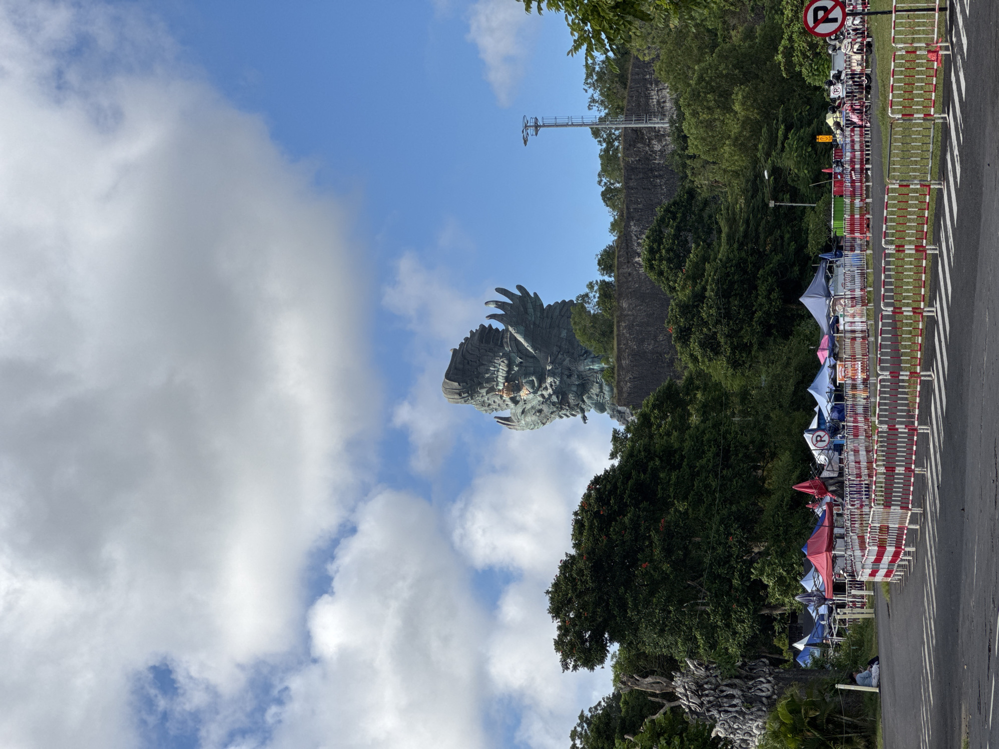 The image, oriented vertically, presents a vibrant outdoor scene, likely at the Garuda Wisnu Kencana Cultural Park in Kuta Selatan, Indonesia. Dominating the upper portion of the frame is a monumental, greyish-green statue of a mythical bird-like figure, resembling Garuda, with intricate detailing visible on its head and wings. It stands majestically against a backdrop of a clear blue sky interspersed with fluffy white cumulus clouds.
Beneath and to the right of the statue, a rugged, dark rock formation or cliff face stretches horizontally across the mid-ground, densely covered in lush green trees and tropical foliage. A tall, slender metal utility pole or tower rises from behind this cliff into the sky.
In the lower part of the image, a paved road with distinct white zebra crossings is visible, running alongside a long array of red and white striped pedestrian barriers. Behind these barriers, a series of small, colorful tents or market stalls, primarily blue, red, and white, are set up. Several people can be seen near these stalls, and numerous motorbikes are parked in the background, suggesting a bustling vendor area or gathering. A prominent red circular sign with a white 'P' crossed by a diagonal line, indicating 'No Parking,' is clearly visible above the barriers.
The bright daylight and scattered clouds suggest the photograph was taken during the day, likely in the morning or early afternoon, with clear weather conditions. The overall atmosphere points to a popular tourist destination with active commercial and visitor facilities.
