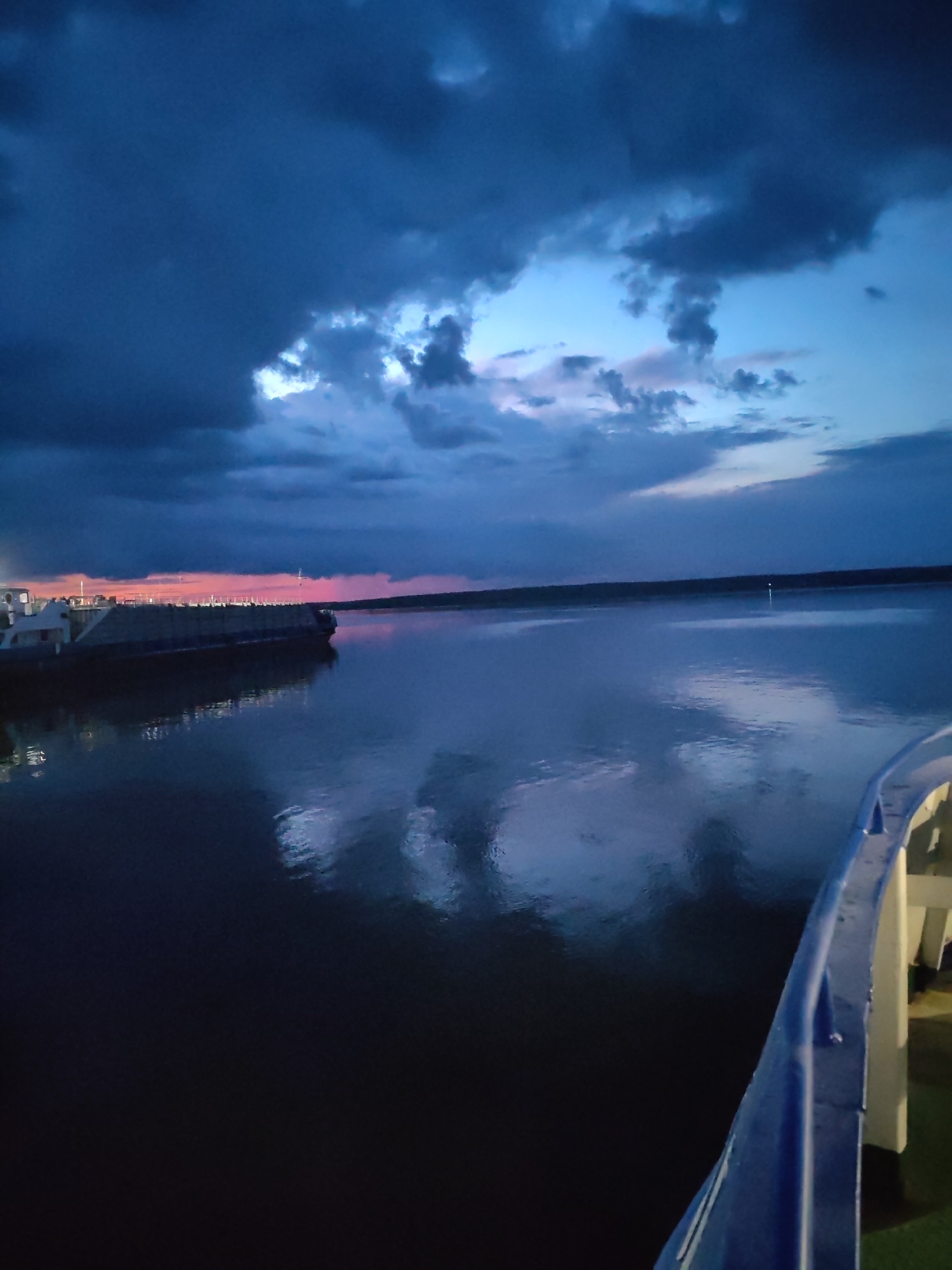 This image depicts a twilight scene on the Volga River near Rybinsk, Russia, as indicated by the context of its potential location. A barge, likely a cargo vessel, is positioned on the left side of the frame, its dark silhouette contrasting with the soft colors of the sunset. The sky above is filled with dramatic, deep blue and grey clouds, with a faint band of pink and orange visible near the horizon, suggesting the last remnants of daylight. The river's surface is calm and reflects the colors of the sky and the barge, creating a mirror-like effect. In the foreground, a blue railing, possibly part of a ship or pier, curves into the frame from the right, indicating the viewer's vantage point is from another vessel or a structure over the water. No people are visible in the scene. The overall atmosphere is serene and tranquil, characteristic of the evening on a large body of water.