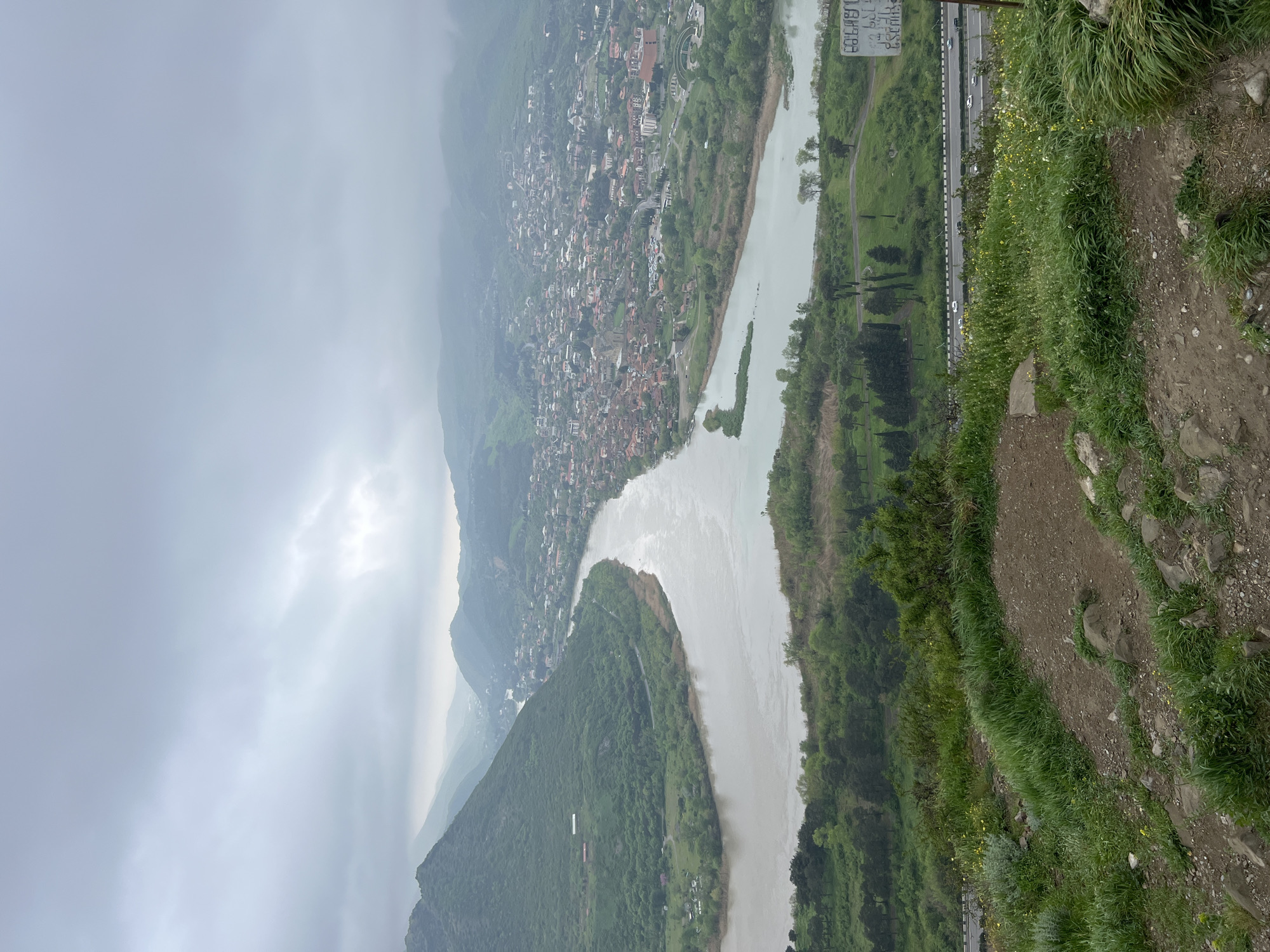 A high-angle, landscape-oriented photograph depicts an overcast day in Mukhatgverdi, Georgia. A winding, light-colored river dominates the scene, curving sharply amidst green, mountainous terrain. A town is nestled on the left side of the river, its buildings appearing dense and close together. On the right side, a highway runs parallel to the river, with vehicles visible on the road. The foreground shows a rocky, grassy slope, suggesting a steep vantage point. The sky is filled with heavy clouds, obscuring the mountain peaks.