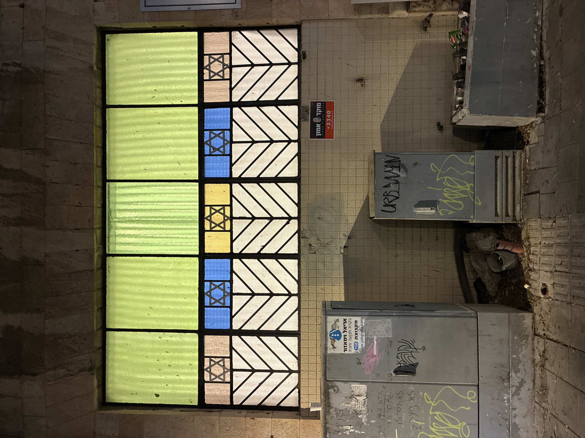 The image depicts a street scene in what appears to be Tel Aviv, Israel. A colorful stained-glass window featuring Stars of David is prominently displayed, indicating a possible religious or cultural significance. To the right of the window, a sign with Hebrew text is affixed to the wall. Below, there are electrical utility boxes covered in graffiti. The surrounding area includes tiled walls, curbs, and some litter. The scene is bathed in artificial light, suggesting it is either nighttime or the entrance to a building.