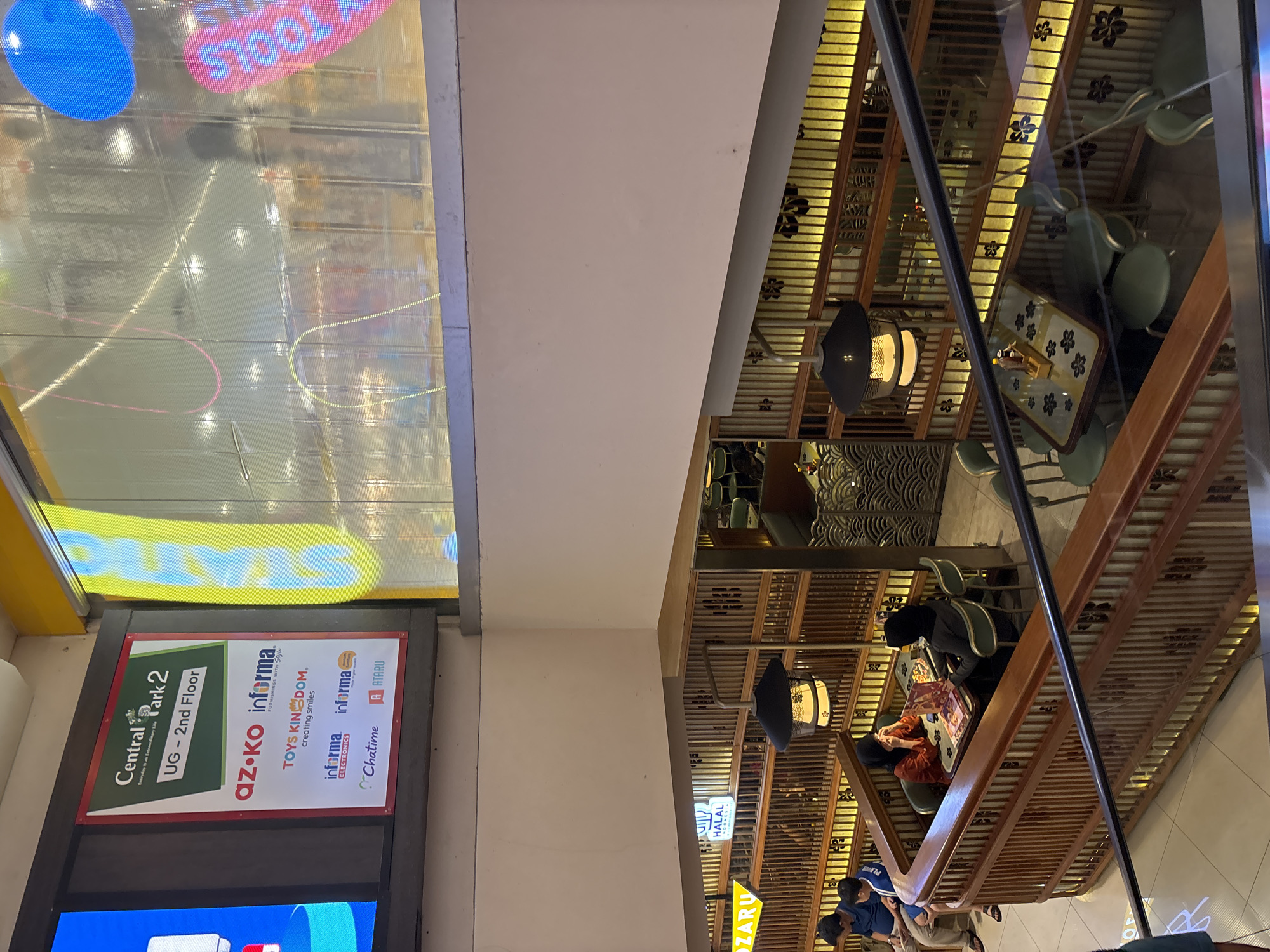 The image shows an indoor scene, likely a mall or shopping center, specifically Central Park 2 in West Jakarta, Indonesia. A sign indicates that the photo was taken on the UG-2nd Floor. The sign also lists several businesses including "az-ko informa," "TOYS KINGDOM," "informa ELECTRONICS," "Chatime," and "A ATARU." To the right of the sign is a ceiling with a complex wooden frame design and Japanese-style hanging lanterns. People are seated at tables, possibly in a restaurant or food court. Some of them are wearing headscarves. A "HALAL" sign is visible. The scene is captured from an elevated perspective.