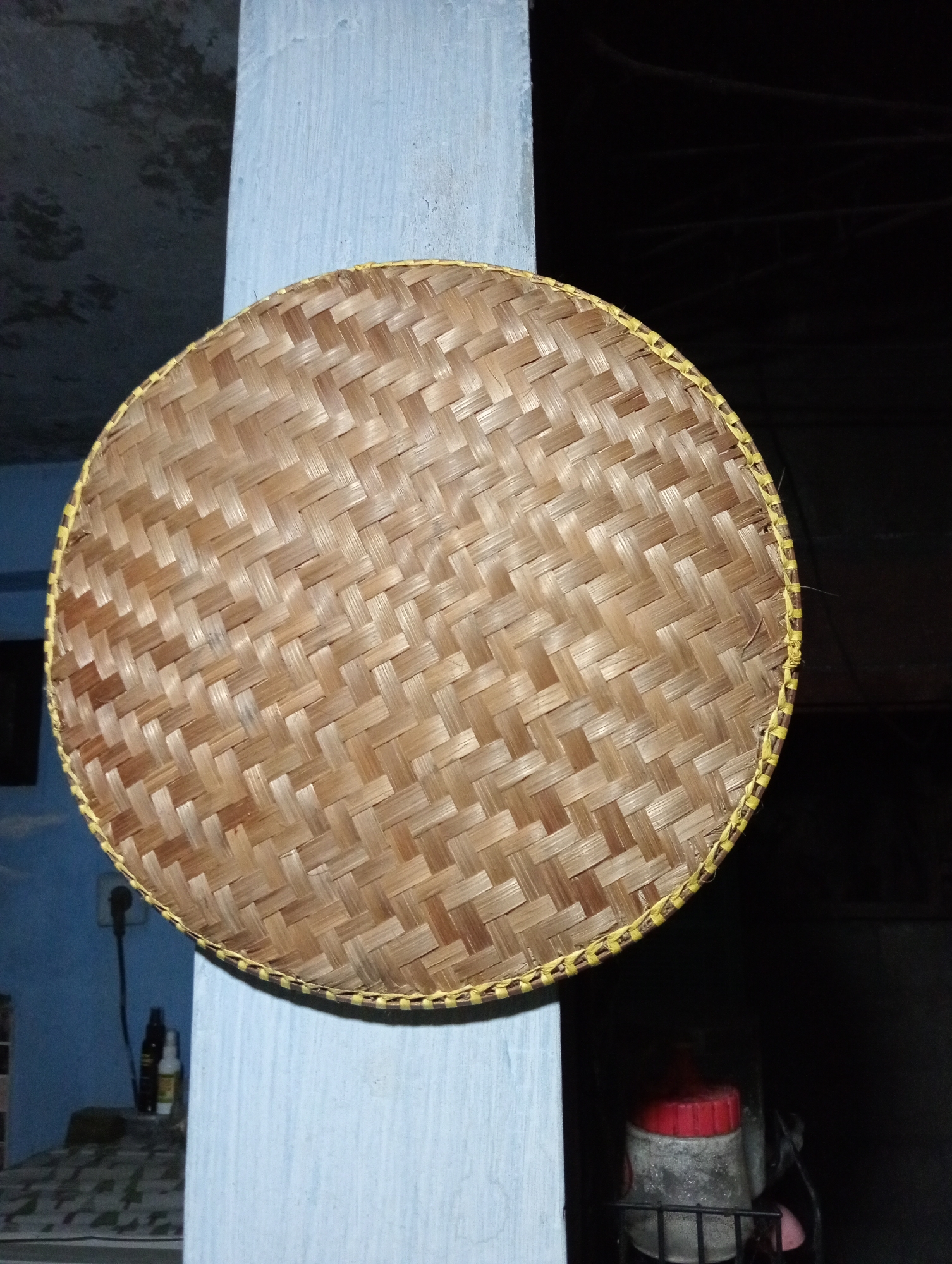 A round, woven bamboo tray with a yellow-stitched edge is centered on a light blue pillar. The tray's surface displays a herringbone pattern. To the left of the pillar, a wall painted light blue is visible, with a table underneath featuring a patterned surface. On the table are two bottles, one dark and one light-colored. A black electrical outlet is mounted on the wall above the table. To the right of the pillar, the background is dark, with a glimpse of a red-lidded container on a shelf. The scene appears to be indoors, possibly in a residence or shop in Trenggalek, Indonesia, given the context.