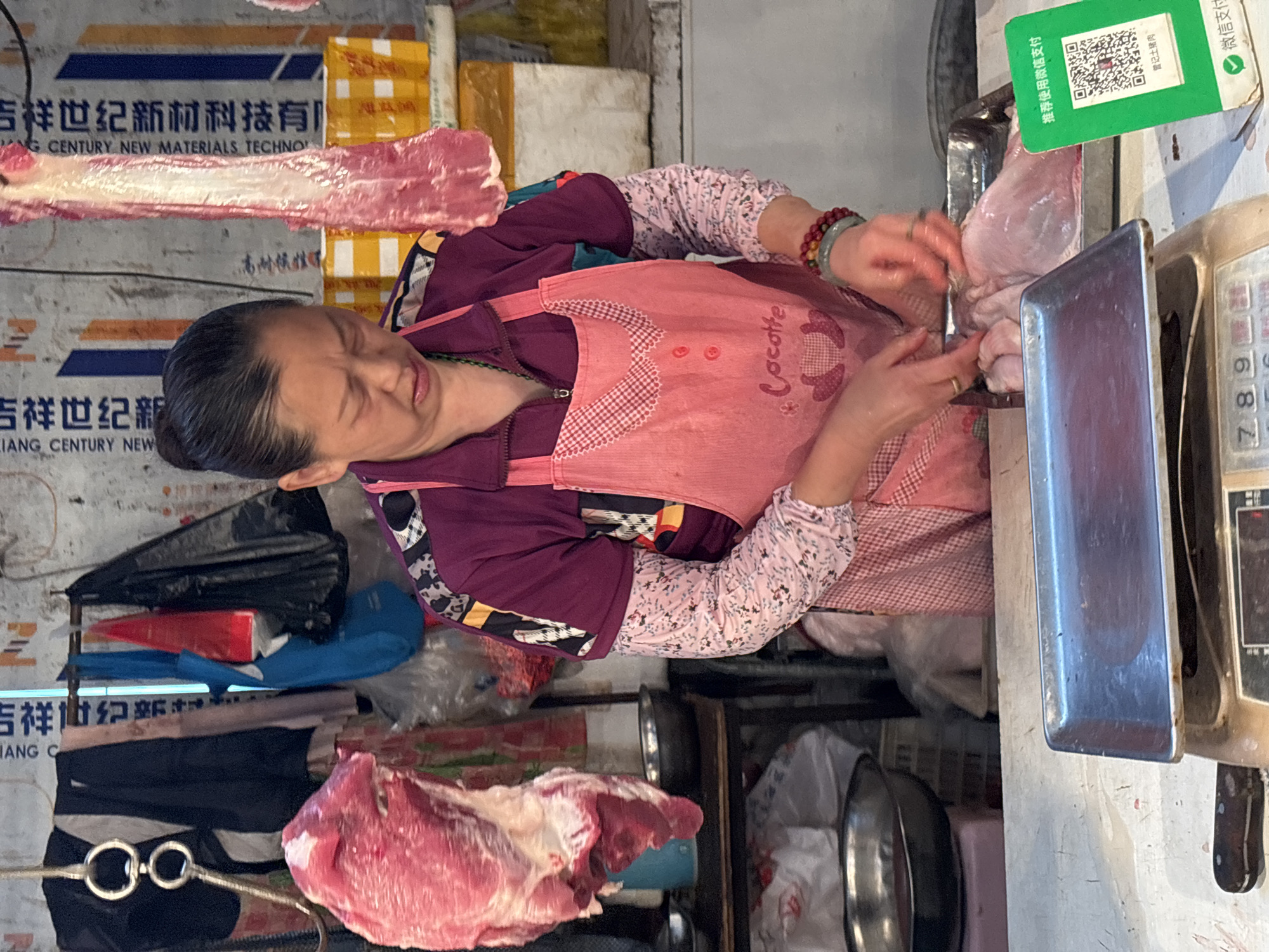 The image shows a middle-aged Asian woman, likely a butcher, working in a market stall in Jiangjin, China. She is wearing a pink apron and floral-patterned sleeves over a purple jacket. She appears to be cutting meat at a counter with a cleaver or knife. A scale is beside her, along with a QR code payment sign for WeChat, indicating a modern payment method is accepted. Various cuts of raw pork are visible, both on the counter and hanging above. Background details include signs with Chinese characters and English text reading "JIANG CENTURY NEW MATERIALS TECHNOLOGY." There are also miscellaneous items such as bowls, plastic bags, and a folded black umbrella visible in the background.