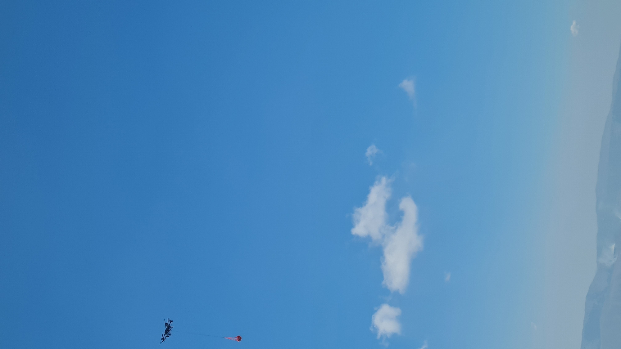 The image shows a clear blue sky with a few scattered white clouds. In the upper left quadrant, a black drone is visible, hovering in the air. A small, red object, possibly a bucket or container, is suspended from the drone by a thin line. The drone and its payload are positioned against the vast expanse of the sky. In the distance, at the bottom of the image, a hazy, mountainous landscape is discernible, with faint plumes of smoke rising from the terrain, suggesting a potential wildfire or controlled burn in the L'Aquila, Italy area. The overall scene conveys a sense of aerial observation or intervention.