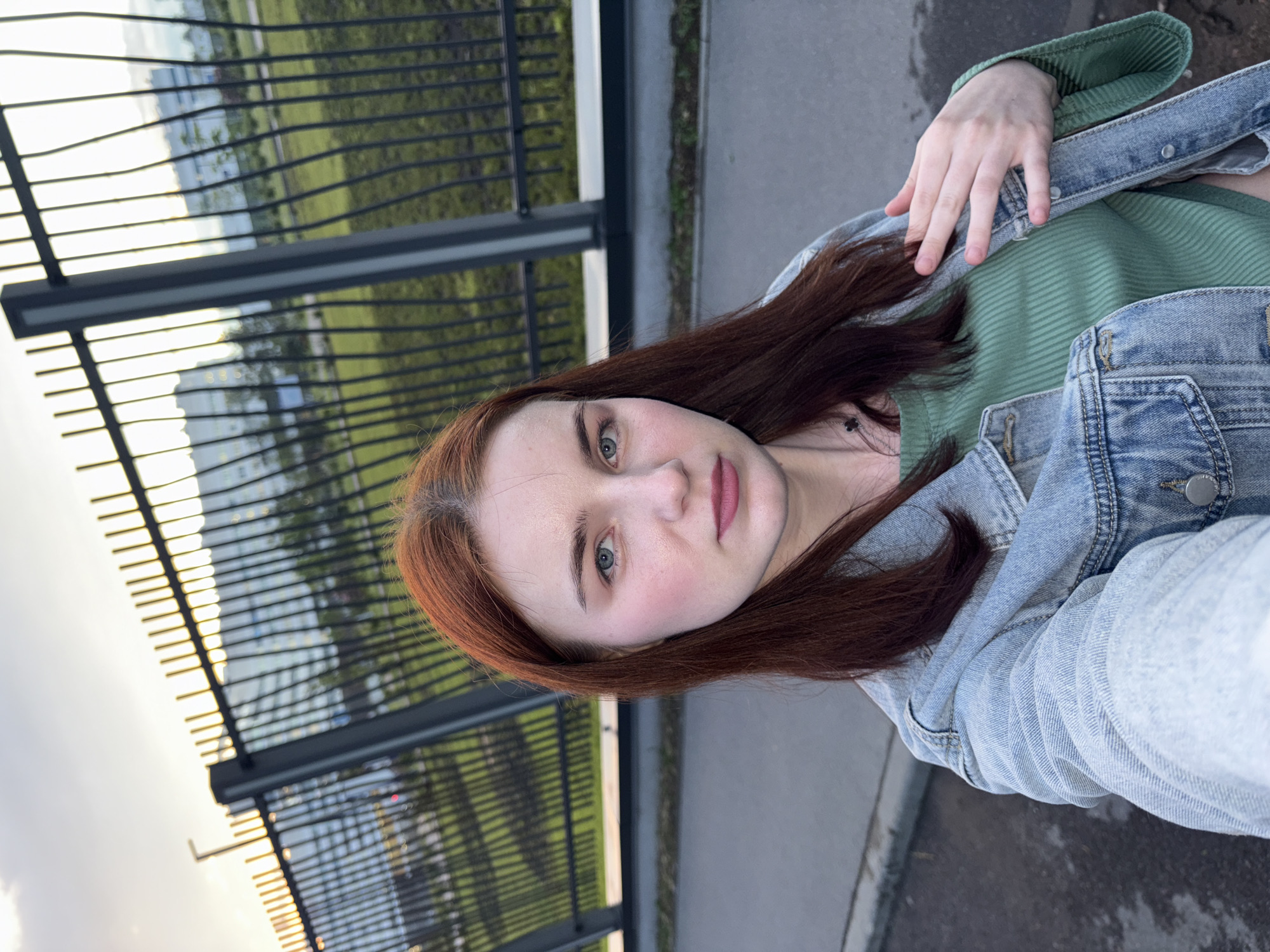 A young woman with auburn hair and blue eyes is taking a selfie outdoors. She is wearing a light blue denim jacket over a green ribbed top. Her left hand is resting on her chest. Behind her, a black metal fence separates her from a grassy area and a modern building. The setting appears to be in the city of Arfa, Russia, during what seems to be late afternoon or early evening, given the lighting. The ground in the foreground is paved.