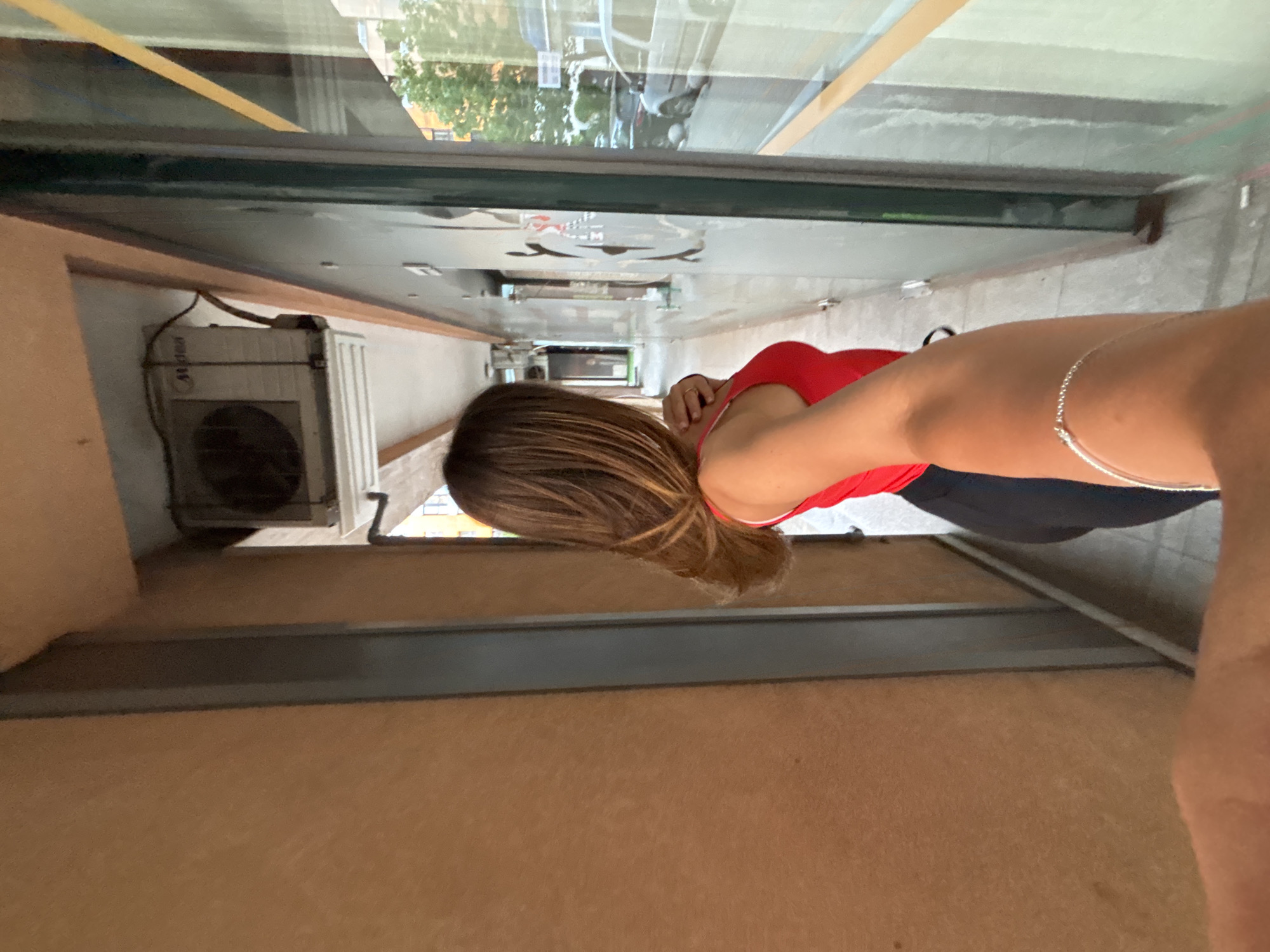 The image shows an indoor scene in Sofia, Bulgaria, as suggested by the reflection in the window. A woman with shoulder-length brown hair and wearing a red tank top and dark pants is partially visible, extending her arm towards the viewer. A silver bracelet is on her wrist. To the left, an air conditioning unit is attached to a wall. The interior walls are painted in a light brown color. Through the reflection in the window, there is a partial view of an urban street setting in daylight.