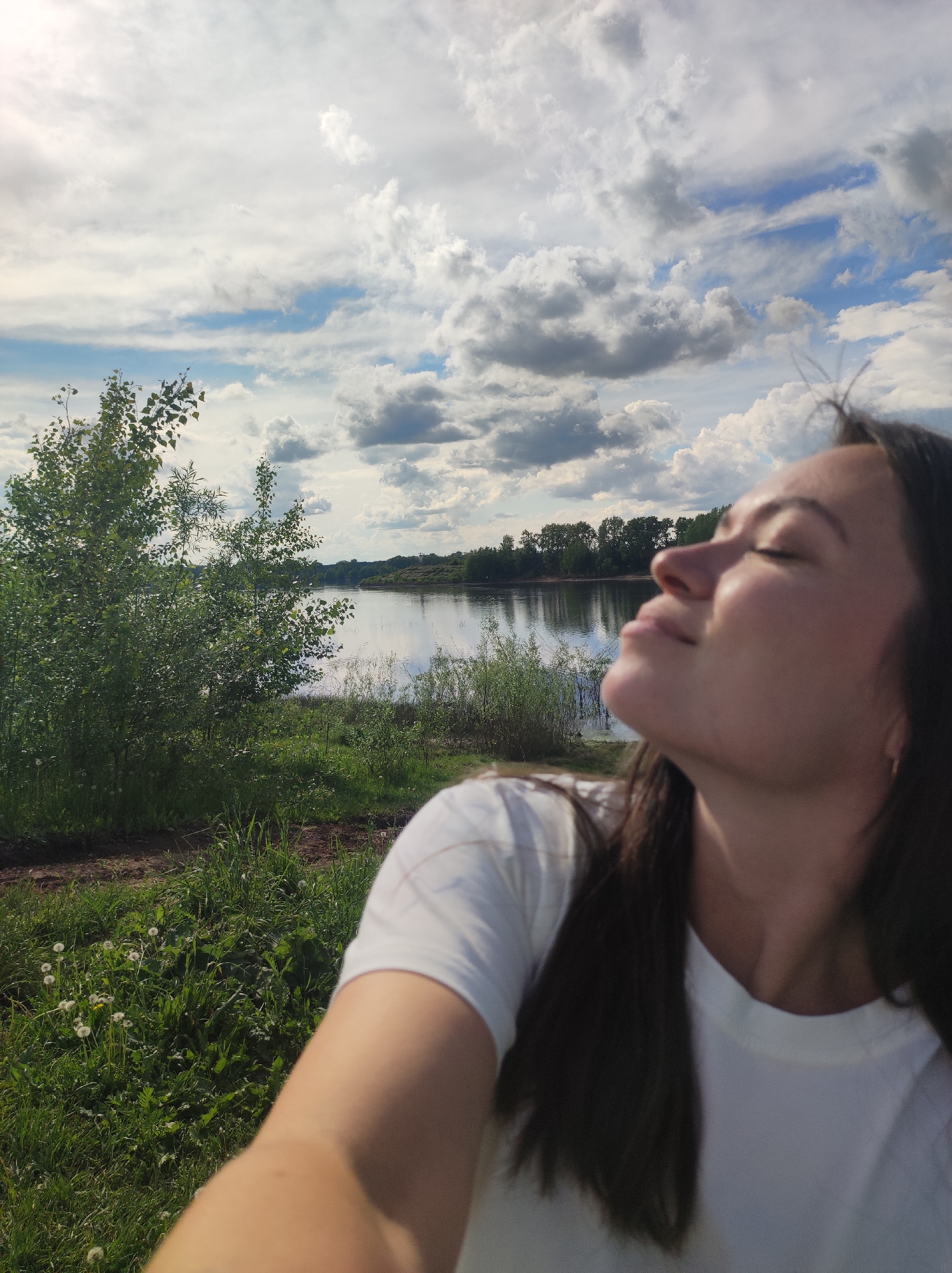 A selfie captures a woman with her face tilted towards the sky. Her eyes are closed, and she wears a white T-shirt. The background reveals a body of water, possibly a lake or wide river, with trees lining the opposite shore. The foreground includes green grass and a young tree with light green leaves. Overhead, the sky is filled with a mix of white and grey clouds against patches of blue. Based on similar images, this is the city of Chaikovsky, Russia.