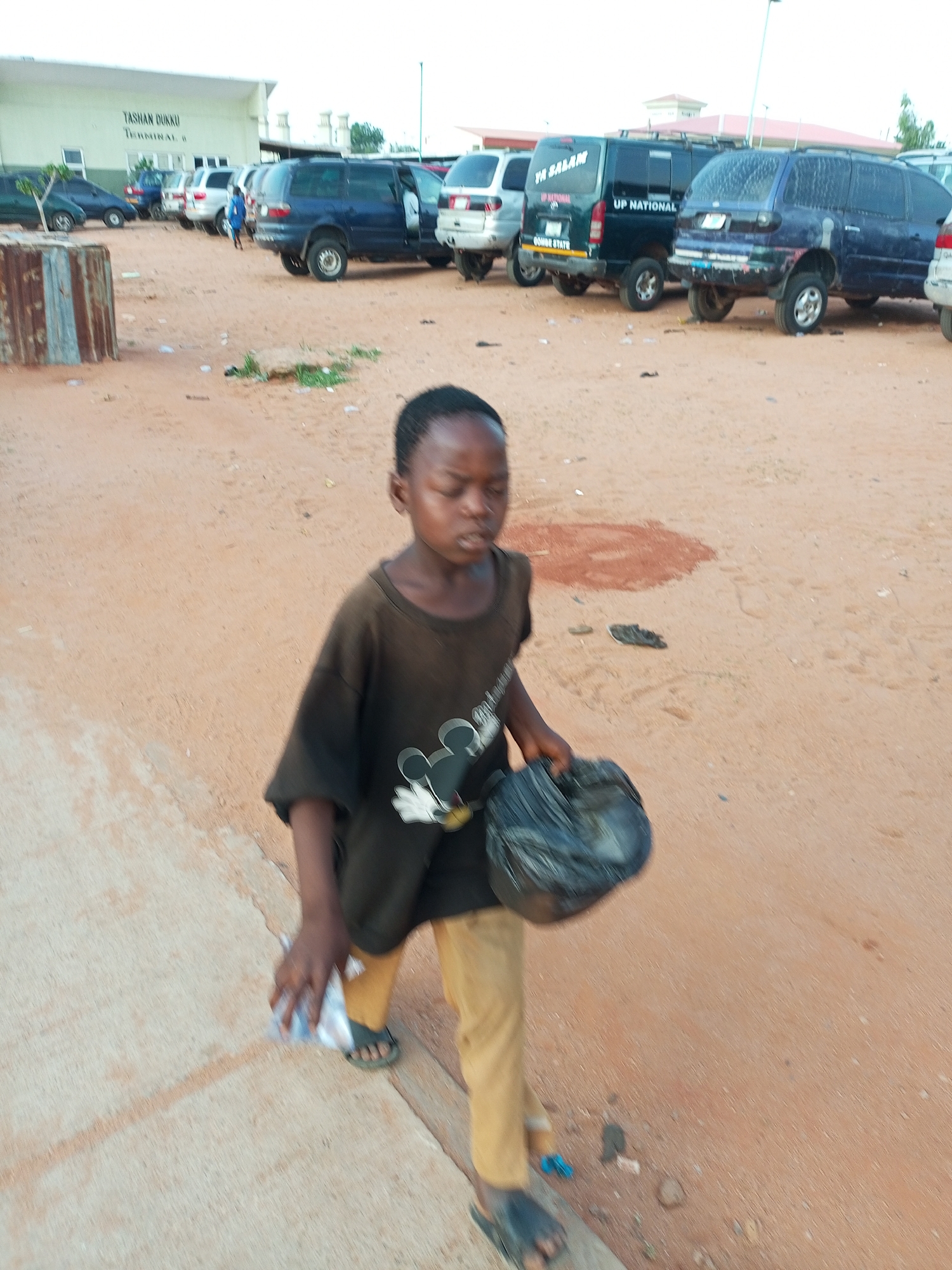 The media file shows a young boy walking on a sandy, dirt ground in a location that appears to be a terminal or transport hub, possibly a bus station. The boy is wearing a dark brown t-shirt with a Mickey Mouse graphic and tan pants. He is carrying a black plastic bag in his right hand. In his left hand, he is holding a clear plastic bag with some items inside. His facial expression is neutral, and he appears to be walking forward.
The background reveals several parked vans and cars, suggesting a busy area. One van has "UP NATIONAL GOMBE STATE" written on it. Further in the background, a building with the sign "TASHAN DUKKU TERMINAL 6" is visible, indicating the location as Gombe, Nigeria. The sky appears overcast or it is late in the day, as the lighting is subdued. The ground is dry and dusty, with scattered debris. The scene is candid and appears to capture a moment in the boy's daily life in this busy transport environment.