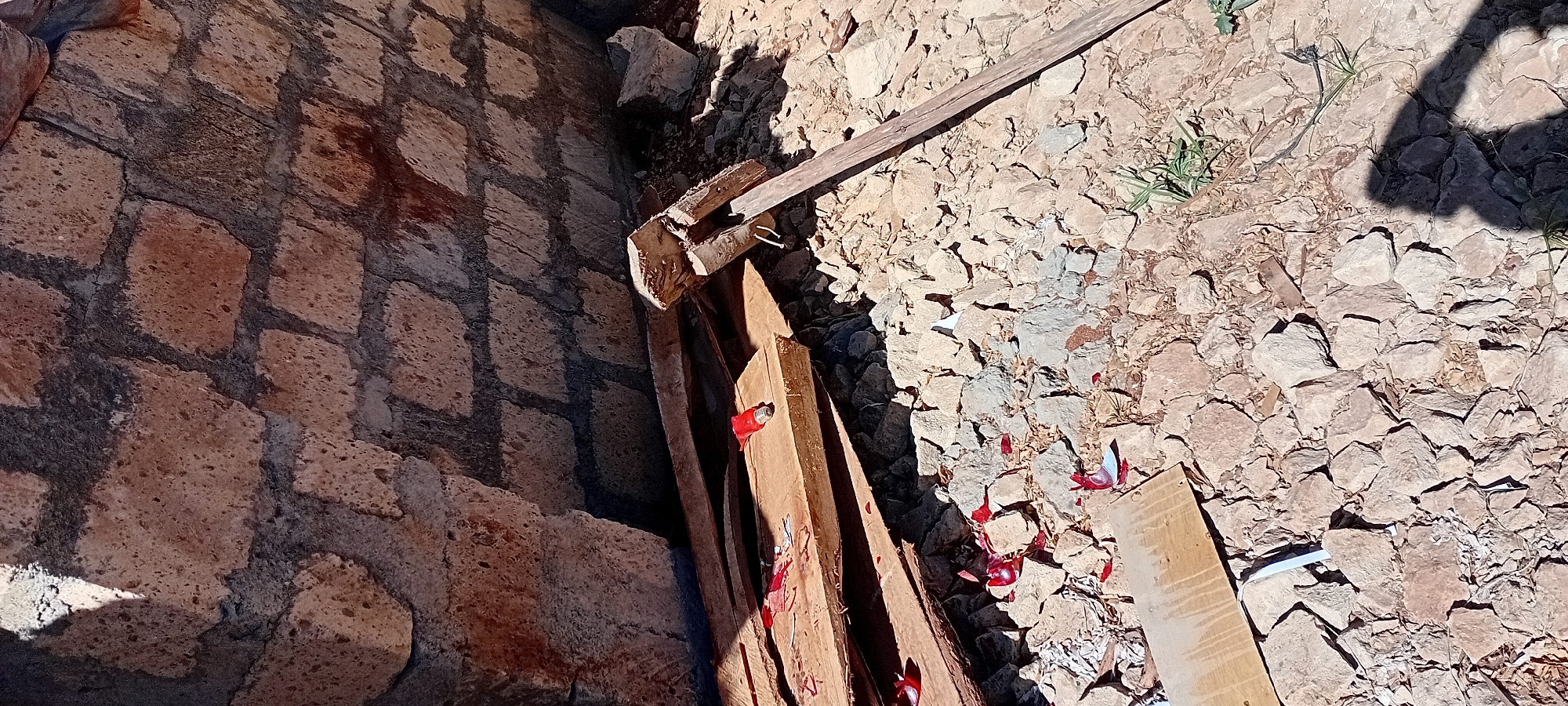 This is a close-up, ground-level, vertical, eye-level shot of a construction debris pile against a rough, stone-built wall. The wall is constructed from irregular, light brown stones mortared together with dark grey cement. The ground is covered with small, light brown stones, dirt, and some dry vegetation. In the center of the frame, a collection of wooden planks is arranged horizontally. These planks appear to be part of construction material, with some showing signs of damage or wear. On the top plank, a small, red object resembling a lipstick or a marker is visible, along with scattered red shards, possibly from broken glass or plastic. To the right of the planks, a wooden tool with a metal attachment, possibly a hammer or a hoe, rests on the ground. The scene suggests an outdoor setting, likely a construction site or an area where building materials and debris have been accumulated. The lighting indicates it is daytime, with shadows cast on the ground and wall. The specific location within the Kijabe ward, Kenya, is not visually identifiable from the provided image.