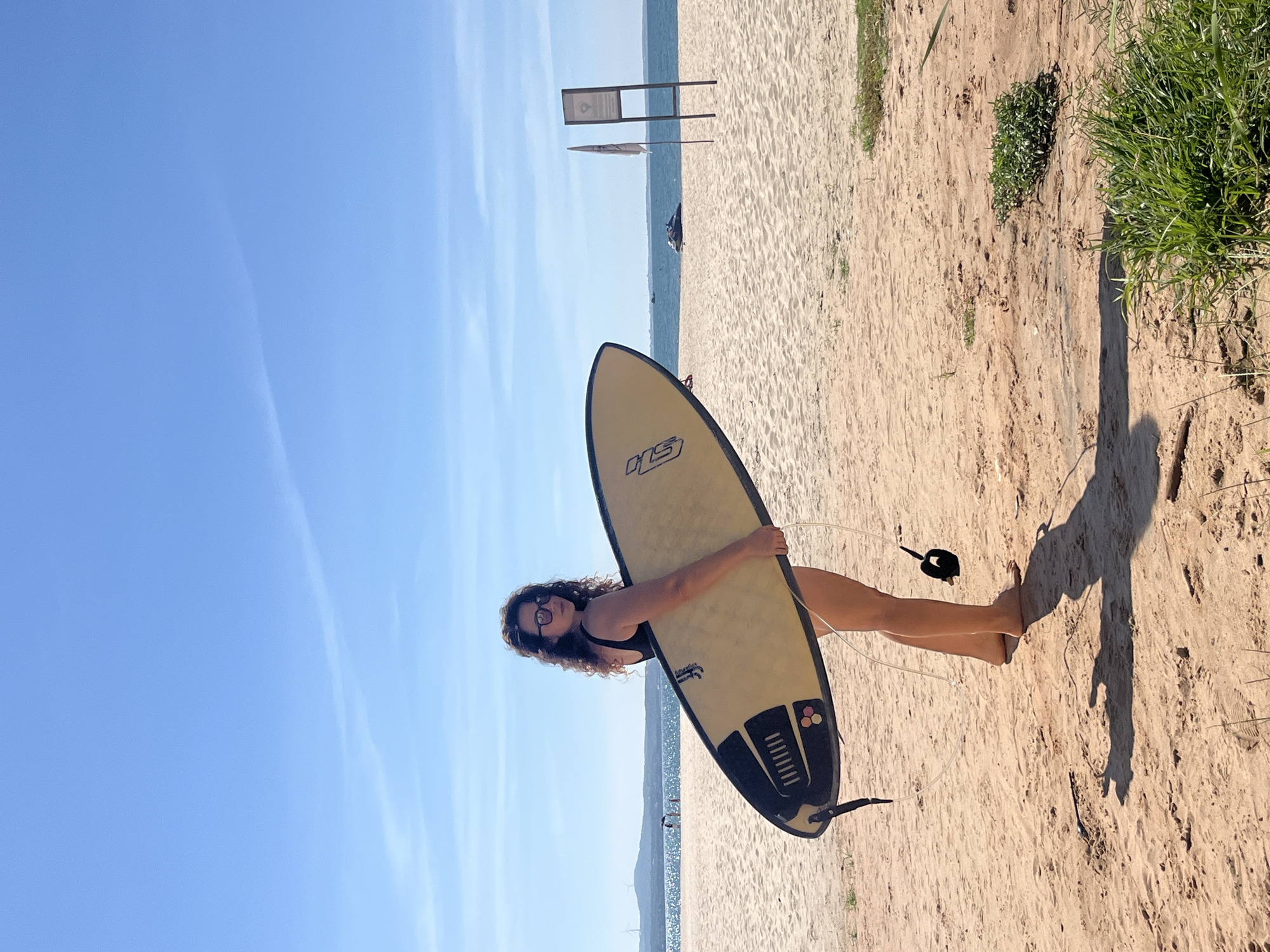 A woman stands on a sandy beach in Phu Thuy Ward, Vietnam, holding a surfboard. She is wearing a black swimsuit and glasses, with curly brown hair. The surfboard is light yellow with black trim and a black leash. In the background, the ocean meets a clear blue sky with a few wispy clouds. The beach is mostly bare sand, with some sparse vegetation and a few distant figures. A signpost and a flag are visible on the beach near the water's edge. A jet ski is in the water in the distance. The lighting suggests it is daytime, with bright sunlight casting shadows.