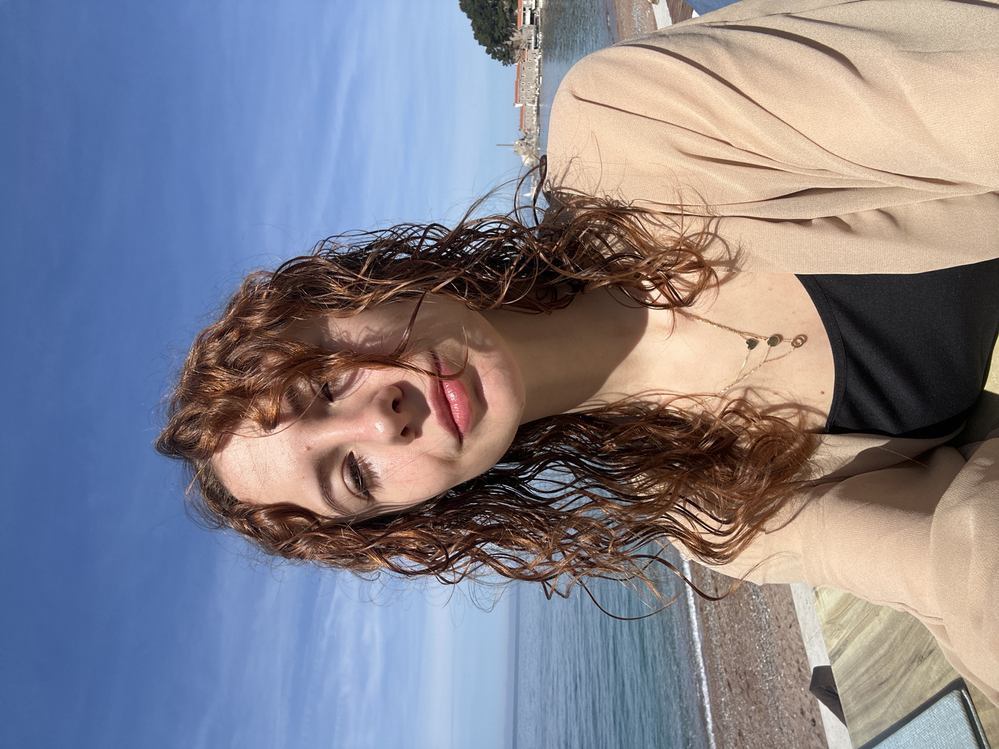A young woman with red, curly, damp hair is shown in a selfie. She is wearing a black top and a tan-colored jacket or sweater. A delicate gold necklace with small charms is visible around her neck. The background consists of a clear blue sky, a body of water (likely the Adriatic Sea), and a distant cityscape that resembles the Petrovac centar area in Montenegro. A small beach is visible at the bottom of the frame, with a boardwalk area. The photograph has been rotated, with the horizon line running vertically across the image.