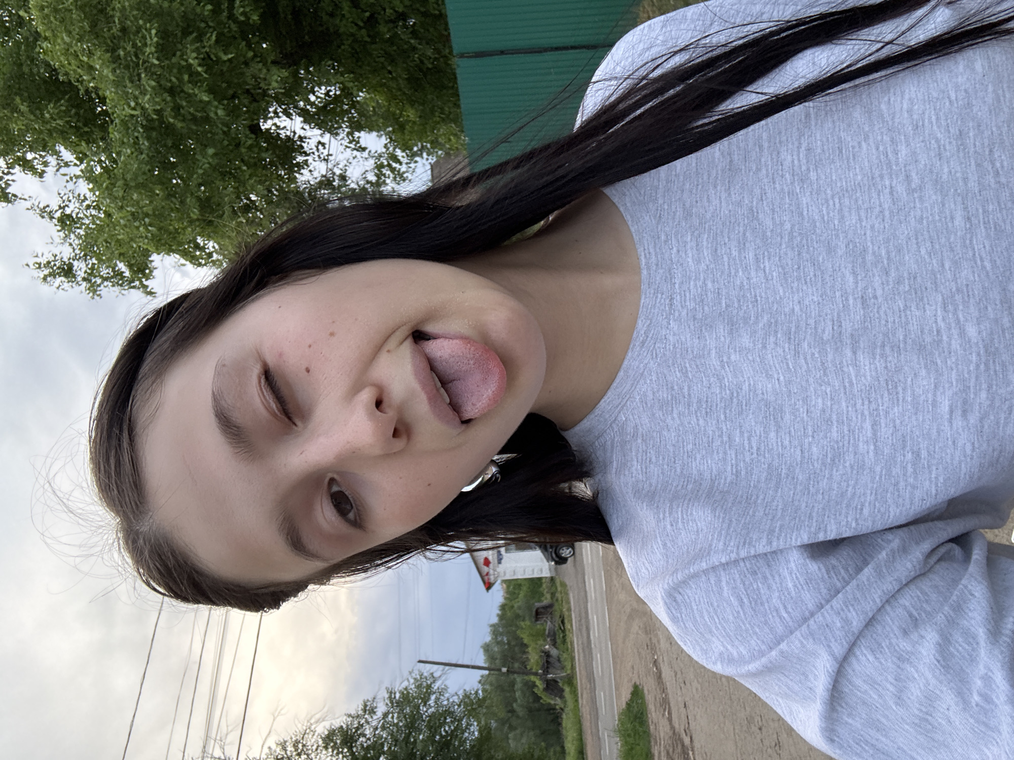 The image shows a young woman outdoors. She has dark hair and is wearing a light gray shirt. She is sticking out her tongue and winking. The background includes trees, a green fence, a road, and a house. Based on the presence of a house with red decorations and a road, the location appears to be in a residential area on the outskirts of Фоки, Russia.