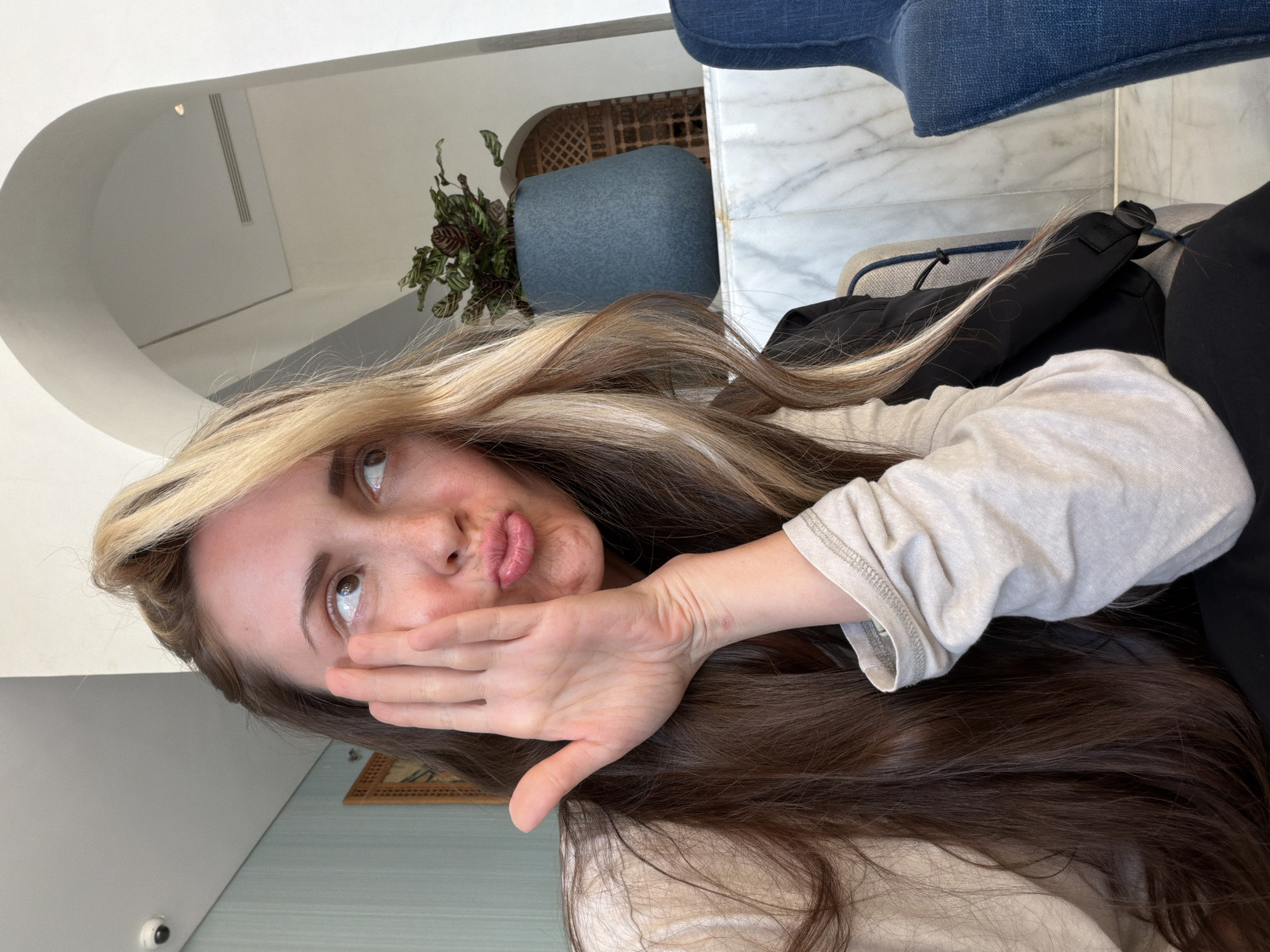 A young woman is featured in an indoor setting, likely a lounge or lobby in Sharm El Sheikh, Egypt. She has long, flowing brown hair with prominent blonde highlights, and light skin with visible freckles across her nose and cheeks. Her brown eyes are directed upwards and slightly to the right, and her lips are pursed in a "duck face" or pouting expression. Her left hand is raised, covering her mouth and part of her cheek. She wears a light-colored (possibly off-white or beige) long-sleeved top and dark outerwear, which appears to be a black jacket.
The background reveals modern interior design elements. On the left, a white arched wall or ceiling feature creates an architectural detail. Further back, a vibrant potted plant with dark green and reddish-purple leaves sits beside a blue upholstered chair. A decorative wooden lattice screen is visible behind the chair. To the right, a white marble table or counter is present, along with a portion of another blue upholstered seating arrangement. The lighting is bright and indirect, suggesting it is daytime. No visible text is discernible in the image. The overall impression is one of a casual, relaxed moment.