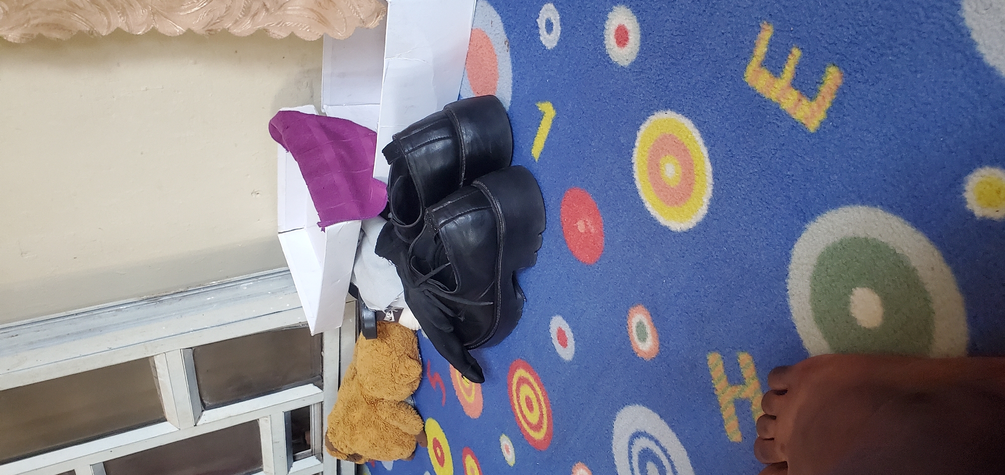 The image shows a pair of black platform shoes with laces, placed on a blue carpet with colorful circular patterns and yellow letter shapes. A small, fluffy brown teddy bear is visible next to the shoes, partially obscured by a purple piece of fabric and white cardboard. A person's foot is also visible in the lower right corner of the image, with dark skin, resting on the carpet. The background includes a cream-colored wall and a window with a metal frame. The lighting suggests it could be daytime, indoors. The overall scene appears to be in a casual setting, possibly a home. There is no visible text in the image.