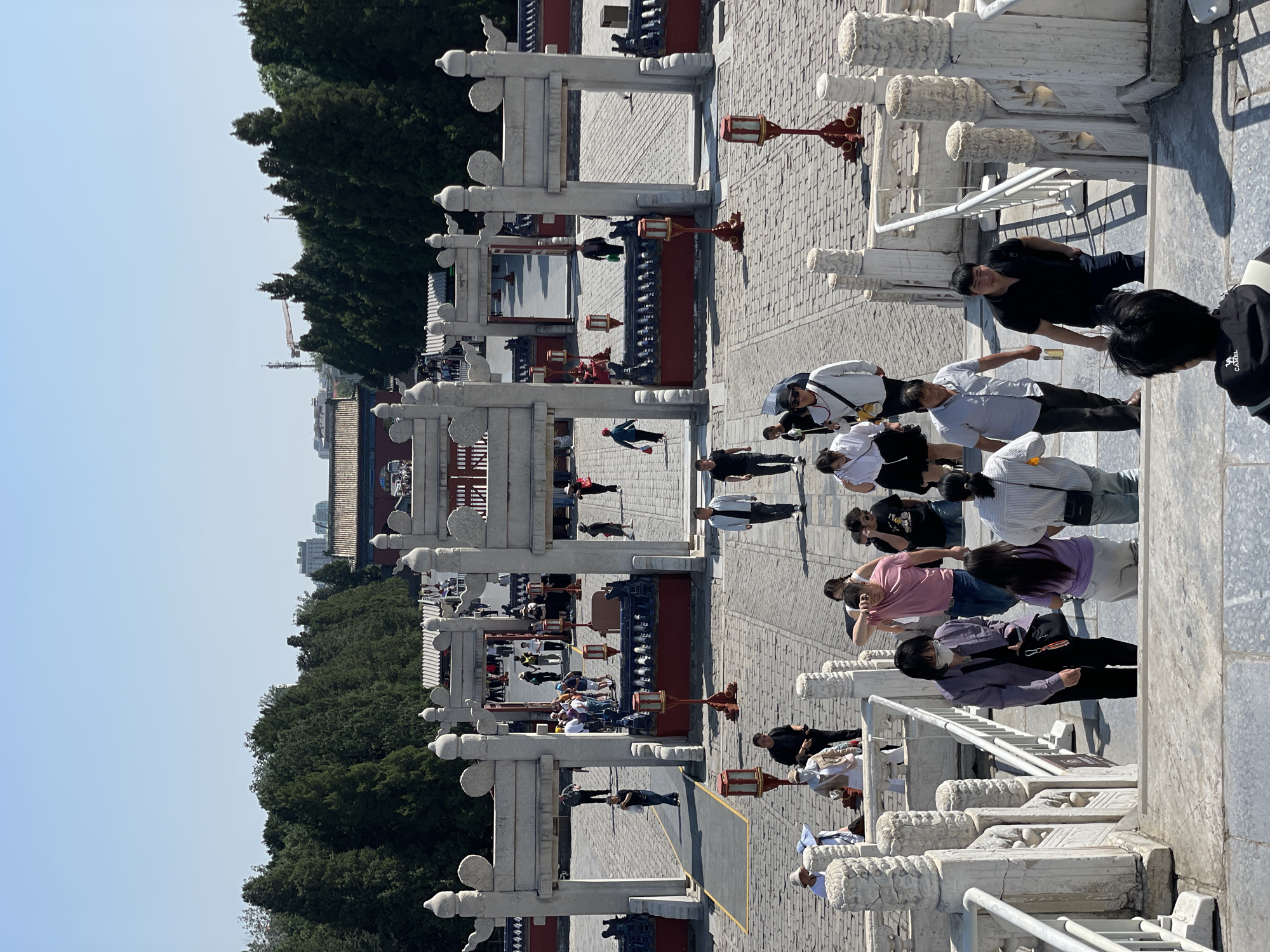 Here is a description of the image:
The image depicts an outdoor scene in Dongcheng District, China. The location is identifiable as the Temple of Heaven based on the distinctive architecture of the gates and pathways. The photograph appears to have been taken from an elevated position, looking down onto a stone pathway.
Several people are present in the scene, seemingly tourists or visitors. They are walking, standing, and interacting with one another. Most are casually dressed in modern clothing.
The pathway is made of stone and is bordered by white stone gates and railings. The gates are elaborately decorated with traditional Chinese designs. Red lanterns are placed at regular intervals along the pathway. Dense foliage from trees lines the pathway and, in the background, a building can be seen, painted red and grey. The sky is a clear blue.