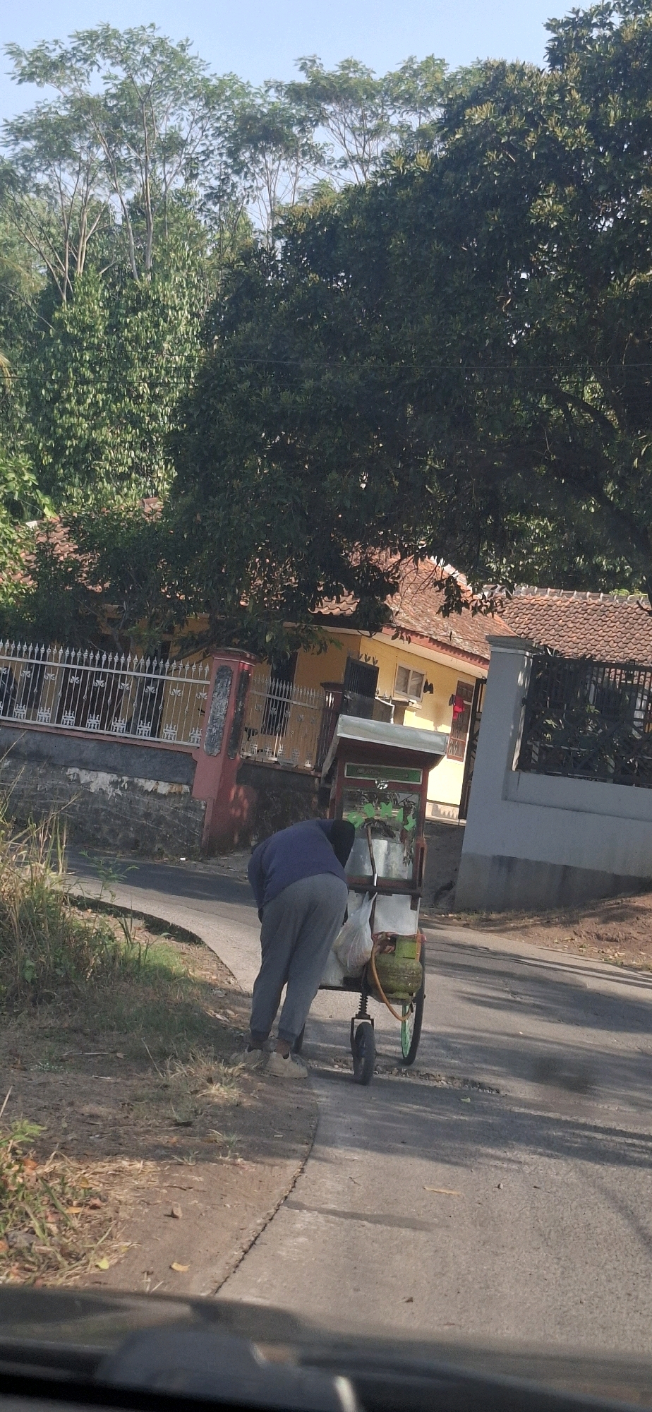 A man wearing a blue long-sleeved shirt and grey pants is bending over to attend to a food cart on the side of a paved road. The cart has a green propane tank attached to its side, and a white plastic bag is hanging from it. The cart has a small canopy and appears to be stocked with goods. The road is surrounded by vegetation, and a house with a red-tiled roof and yellow walls is visible in the background, behind a white metal fence. The sky is clear and blue. This scene is consistent with a street vendor in a semi-rural or residential area of Cibinong Hilir, Indonesia.
