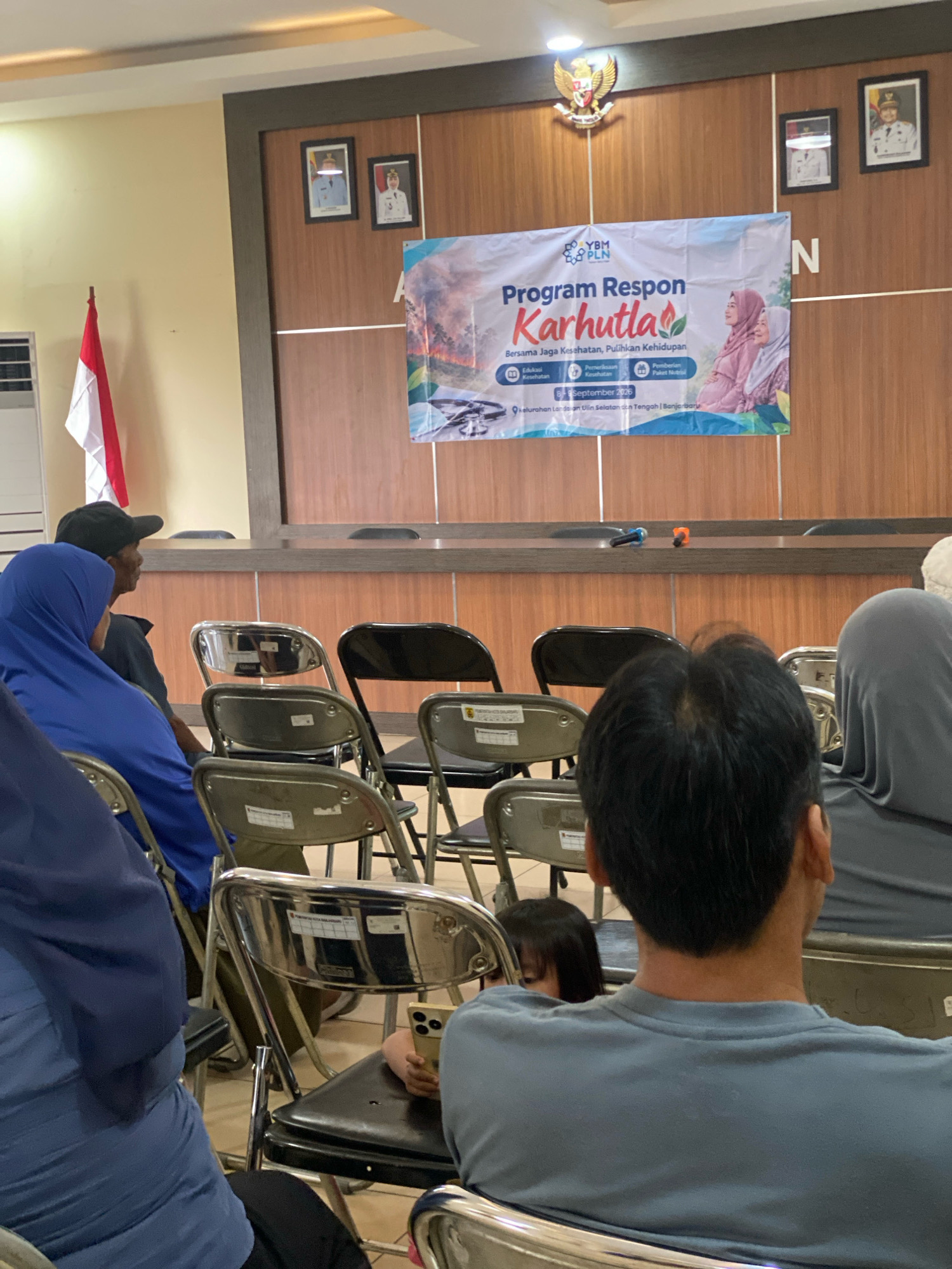 The image depicts an indoor scene, likely a community hall or meeting room, in Banjarbaru, Indonesia. A banner, predominantly white with blue and green accents, is displayed on a wooden paneled wall. The banner announces "Program Respon Karhutla," translating to "Forest and Land Fire Response Program," with the tagline "Bersama Jaga Kesehatan, Pulihkan Kehidupan" ("Together Guard Health, Restore Life"). The banner also includes imagery of a pregnant woman and a child, suggesting a focus on community health and well-being in relation to environmental issues.
In front of the banner, a row of chairs is set up, some occupied by individuals. A man wearing a blue hijab and a person in a black cap are seated on the left. To the right, a man with dark hair and wearing a grey t-shirt is in the foreground, facing away from the camera. A young child is partially visible between two chairs. Microphones are placed on a wooden table in front of the chairs.
On the wall above the banner, two framed portraits of men in uniform are visible, along with a national emblem. To the left of the banner, an Indonesian flag is displayed on a pole. The room appears to be lit by overhead lighting, and the overall atmosphere suggests a public event or seminar. The presence of the flag and the specific banner content indicates a local government or community-organized event related to disaster response and public health.