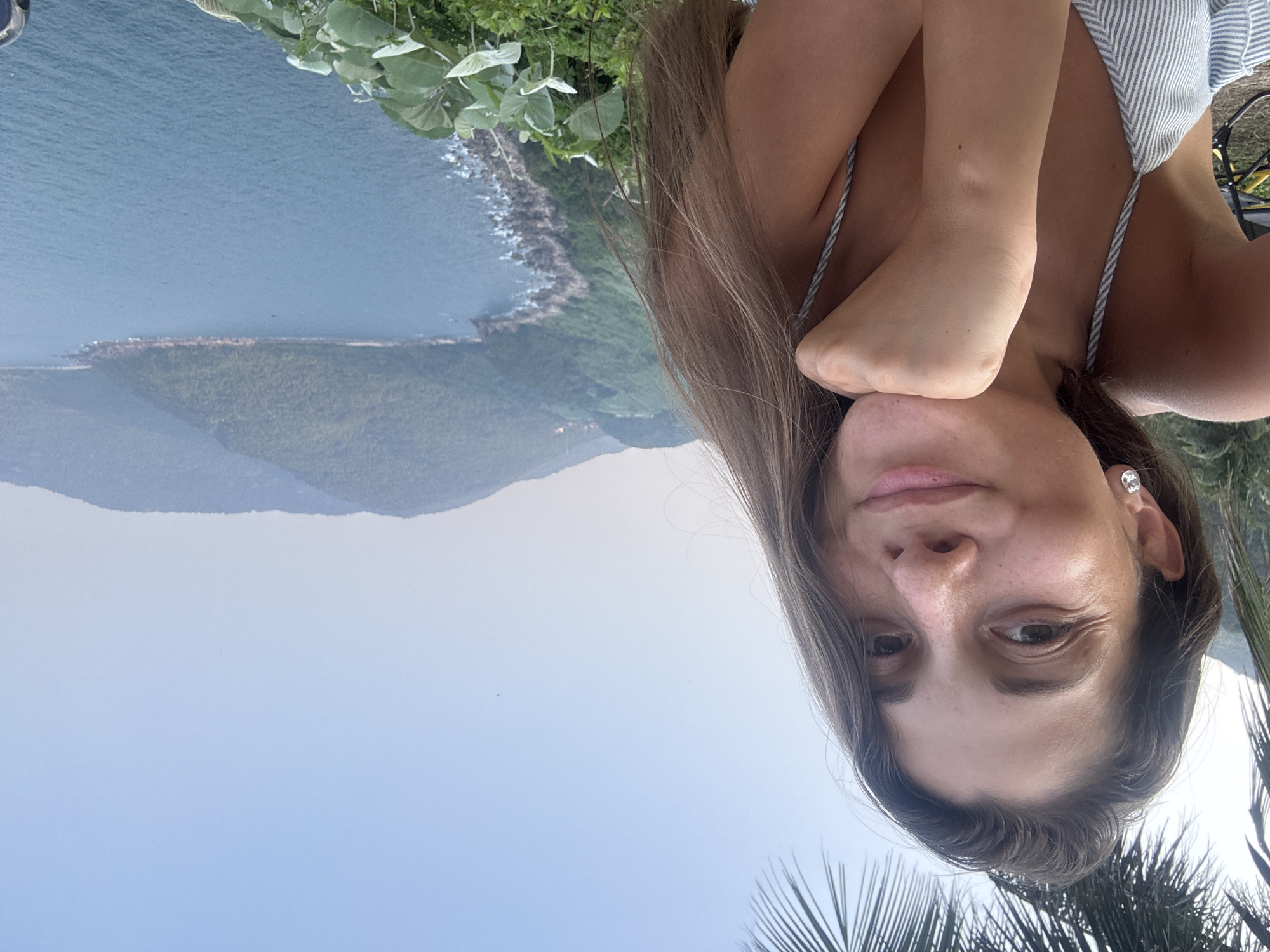 A woman is posing for a selfie at a scenic overlook in Đà Nẵng, Vietnam. She is wearing a striped top and has long brown hair. Her left hand is resting on her chest. In the background, there is a view of the coastline, mountains, and the sea. There are lush green plants surrounding the overlook, including some palm trees in the bottom right corner of the image. The sky is light blue.
