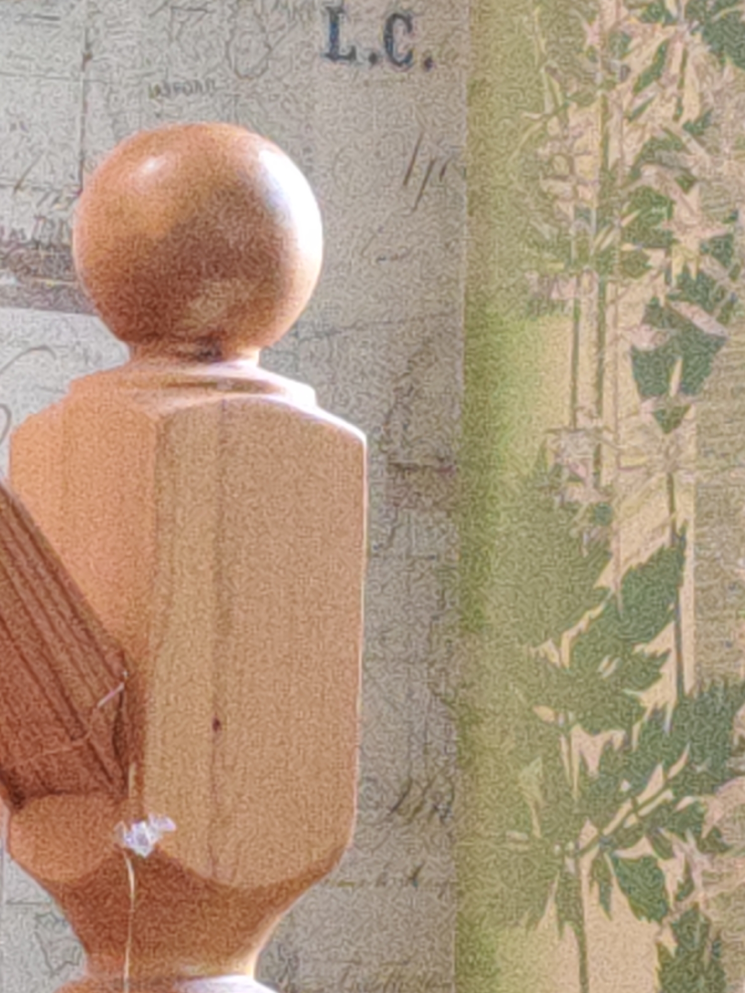 The image shows a close-up of a wooden finial, likely part of a stair railing or furniture. The finial has a spherical top and a faceted base. To the left of the finial, a portion of a wooden spindle with vertical grooves is visible. The background is a textured wallpaper with a faded map-like pattern on the left and a green, leafy design on the right. A small, white, flower-like object is attached to the finial with a thin string or thread. The scene is indoors, with soft lighting. There are no people visible in the image. The location of this object in Rostov-on-Don, Russia cannot be determined from the visual information provided.