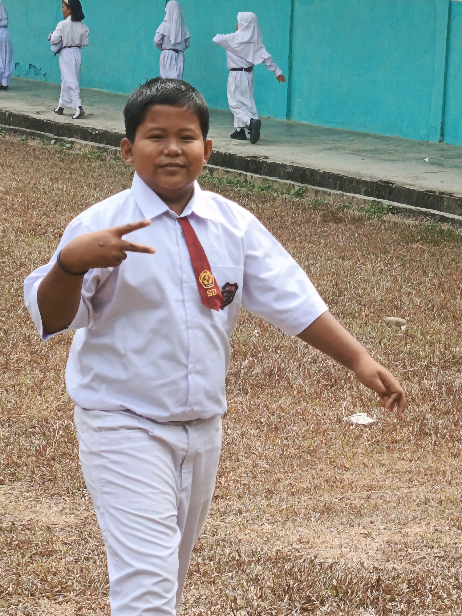 A young boy, dressed in a white school uniform with a red tie and a crest on his shirt pocket, poses for a photo. He is standing on dry, brown grass in an outdoor setting. His left hand is held up, forming a peace sign with his index and middle fingers extended. In the background, several other children in similar white uniforms are visible, some wearing headscarves, standing near a bright turquoise wall. The overall environment suggests a schoolyard in Wonosari, Indonesia, possibly during a school event or activity.