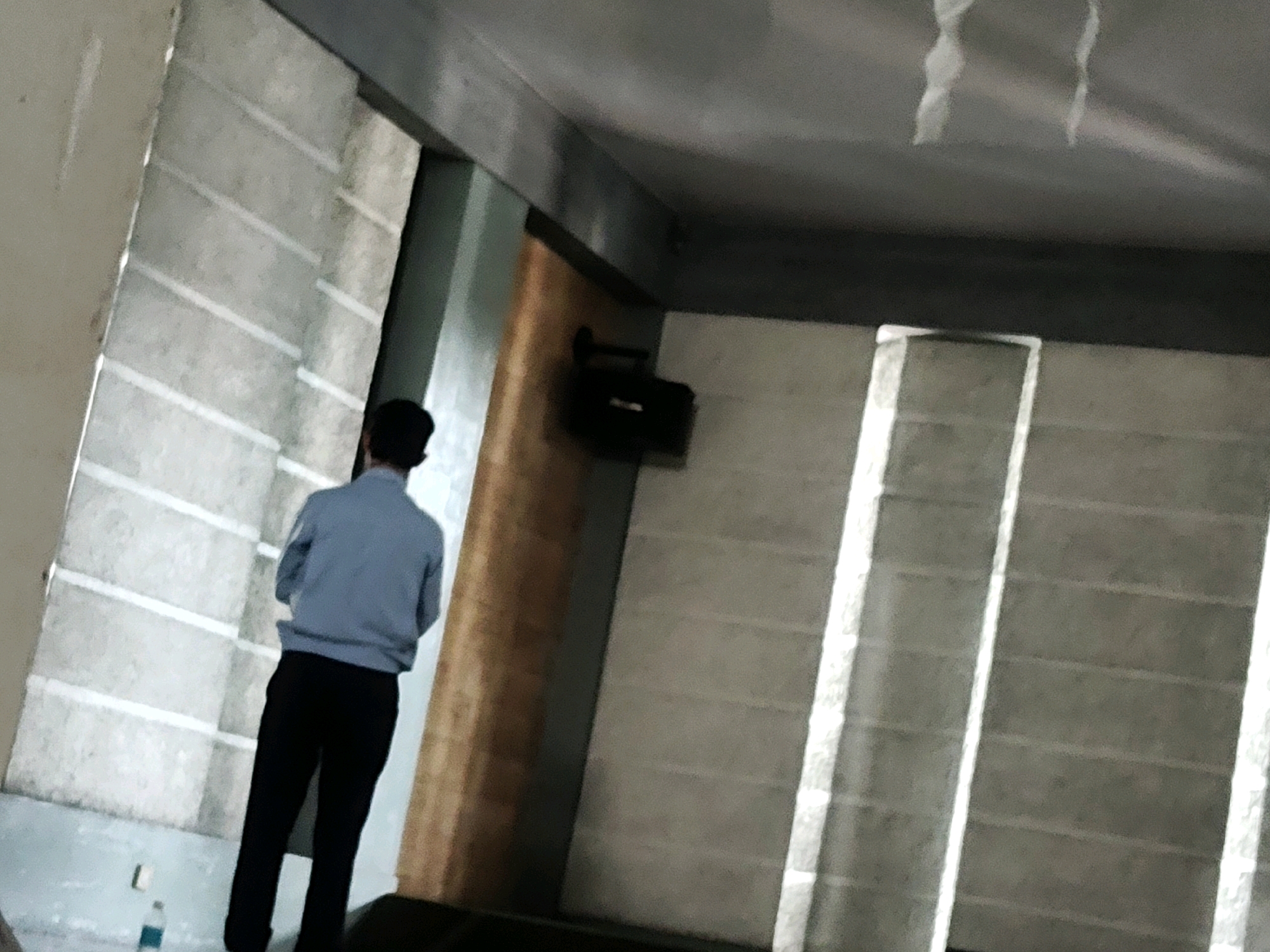 A single individual, appearing to be male and dressed in a light blue long-sleeved shirt and black pants, stands with his back to the camera. He is facing a wall that features horizontal blinds or slats. To his right, a wooden-paneled wall is visible, and further to the right, another wall with similar horizontal blinds. The environment appears to be indoors, with lighting casting bright diagonal stripes across the slatted walls. A dark, box-like object is mounted on the wooden wall above the individual. A small blue bottle lies on the floor near the bottom left of the frame. The overall scene suggests an indoor space, potentially a hall or a room with architectural features designed to manage light. The location is specified as Mekarmukti, Indonesia.