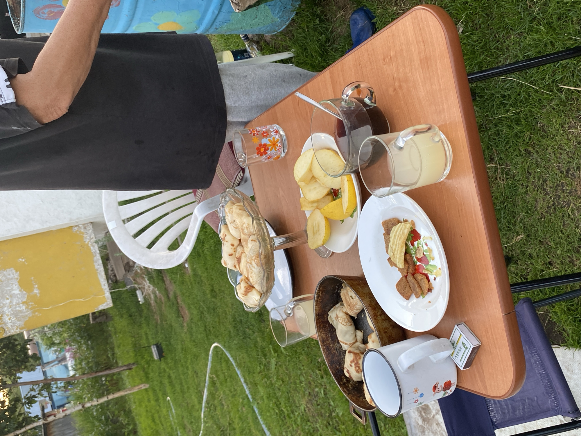 The image depicts an outdoor scene with a table set for a meal. A wooden table is laden with various food items and drinks, including pancakes, sliced fruit, a beverage in a glass mug, and a glass with a yellow liquid, possibly juice. A small box of matches rests on the table next to a mug. In the background, a person wearing a black t-shirt and grey sweatpants stands near a blue barrel with painted flowers. The setting appears to be a yard with green grass and some vegetation. There are no visible signs indicating the specific location in Novyi Ural, Russia.