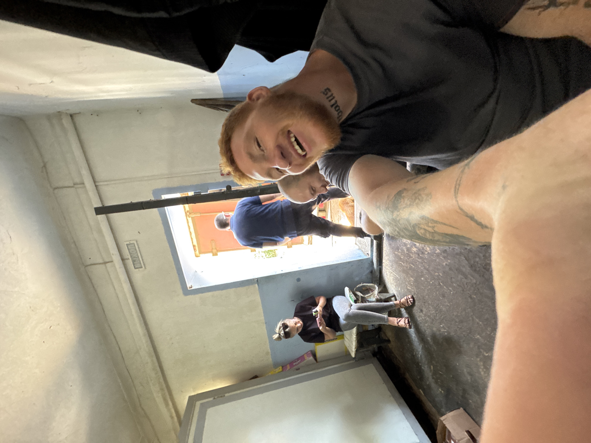 The image depicts an indoor scene with a group of people. In the foreground, a man with red hair and a beard is smiling at the camera, with tattoos visible on his arm and neck. Behind him, another man is partially visible, looking towards the right. To the left, a woman with her hair in a bun is seated on a bench, looking at her phone. She is wearing a black t-shirt, grey jeans, and sandals. In the background, a man in a blue t-shirt and dark pants is walking away from the camera, through an open doorway. The environment appears to be a utilitarian interior, possibly a warehouse or industrial space, with concrete floors and white, blue, and grey walls. There are some boxes and a black garment hanging on the right side of the frame. A sign above the doorway reads "ВЫХОД" (Exit).