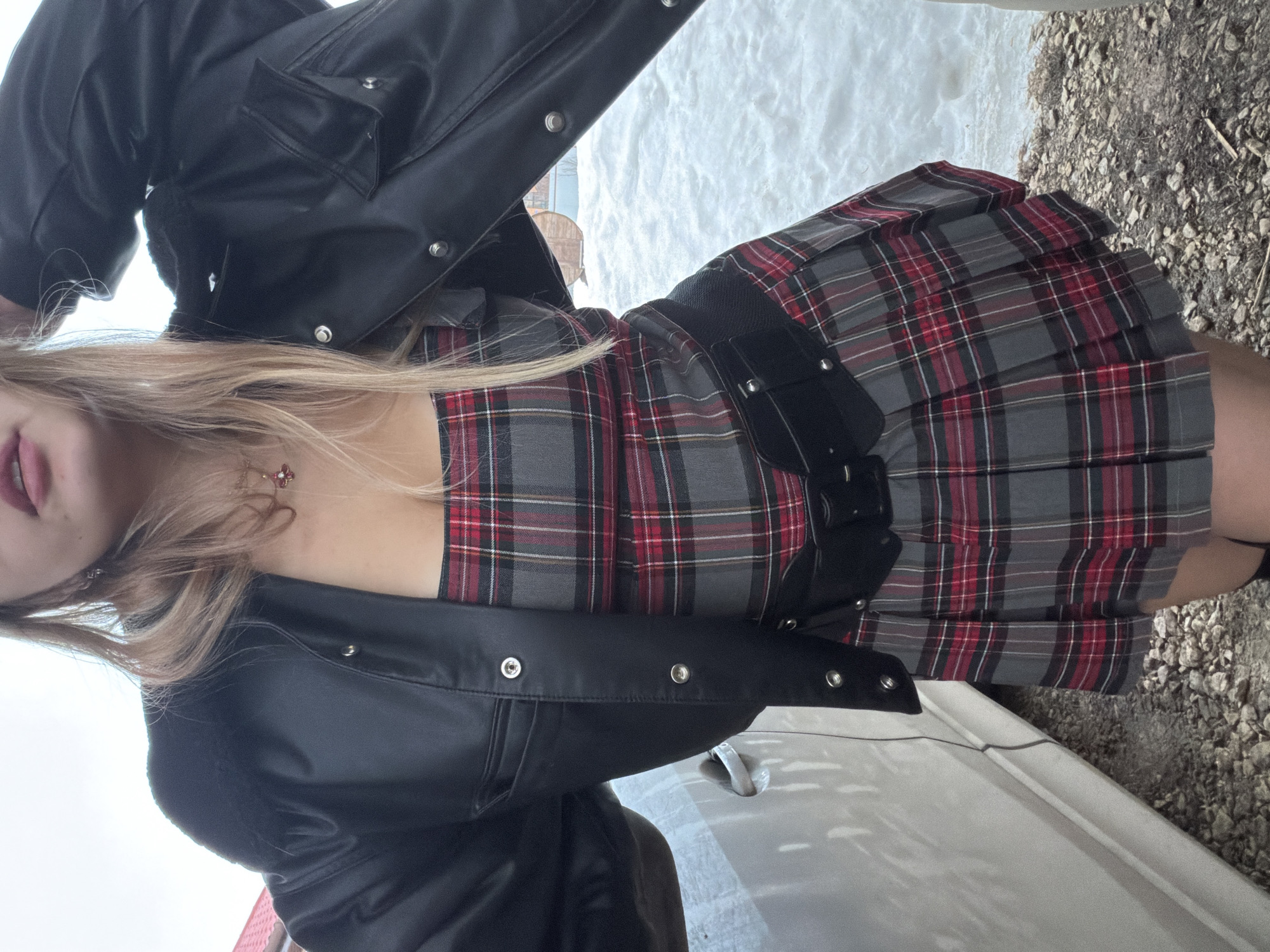 A female subject is depicted outdoors in a candid, waist-up portrait taken in Большая Шильна, Russia. She has long blonde hair partially obscuring her face, with visible pink lips. She wears an unzipped black faux leather bomber jacket over a strapless top or dress featuring a red, grey, and black plaid pattern with a pleated skirt. A wide black belt with multiple metallic fasteners is cinched around her waist. A necklace with a red, flower-shaped pendant rests on her chest. Her left arm is raised with the hand near her head, suggesting a pose. To the immediate left of the subject, the side panel and door handle of a light-colored vehicle, possibly a car, are visible. The ground directly beneath her consists of gravel and dirt. Beyond this, the background on the right features significant snow cover. In the distance, parts of buildings are visible, including a red-tiled roof on the left and a wooden structure on the right. The sky is bright and overcast.