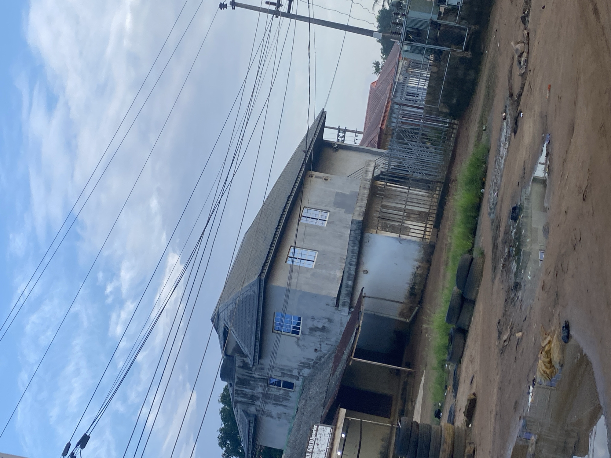 This image depicts a scene in Ikorodu, Nigeria, characterized by a mixture of residential buildings, overhead power lines, and a muddy, somewhat unkempt environment. In the foreground, a dirt road is visible, with puddles of water and scattered debris, suggesting recent rainfall or poor drainage. Several tires are stacked to the left of the frame, and others are scattered on the ground.
Dominating the middle ground is a two-story concrete building with a grey, tiled roof. It has a somewhat weathered facade and features several windows, some with blue-tinted glass. Adjacent to this building, to its left, is a smaller, single-story structure with a corrugated metal roof. Further to the right, another residential building with a red tiled roof is partially visible, separated by a metal gate and fence.
The sky above is partly cloudy with a mix of white clouds against a pale blue background. A prominent feature of the scene is the dense network of overhead power lines crisscrossing the sky and supported by utility poles, indicating the presence of electricity infrastructure. A large electrical transformer is also visible on the right side of the image. The overall impression is that of a residential area in a developing urban or semi-urban setting, where infrastructure is visible alongside signs of everyday life and potentially challenges with waste management and road conditions. No people are clearly visible in the image.