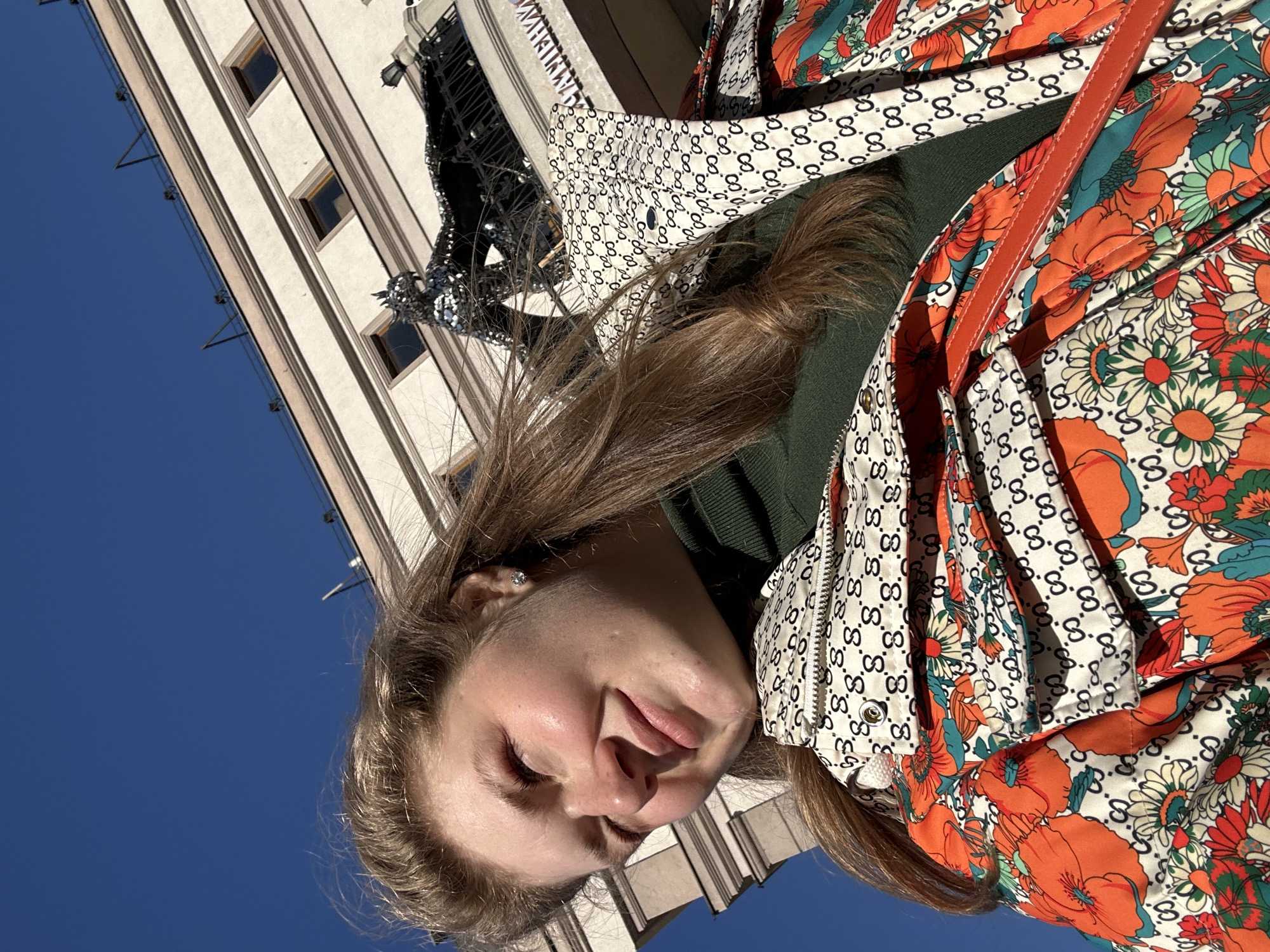 A young woman is depicted from a low-angle perspective, looking down towards the camera, with a partial view of a light-colored building and a section of blue sky in the background. She has fair skin, light brown hair, and is wearing an olive green top under a patterned jacket featuring floral and Gucci-style "G" logo designs. A red strap is visible over her shoulder. The location is suggested to be Kazan, Russia, inferred from visual elements of the architecture or decorative elements on the building, though this detail is not definitively visible in the depicted portion of the image.