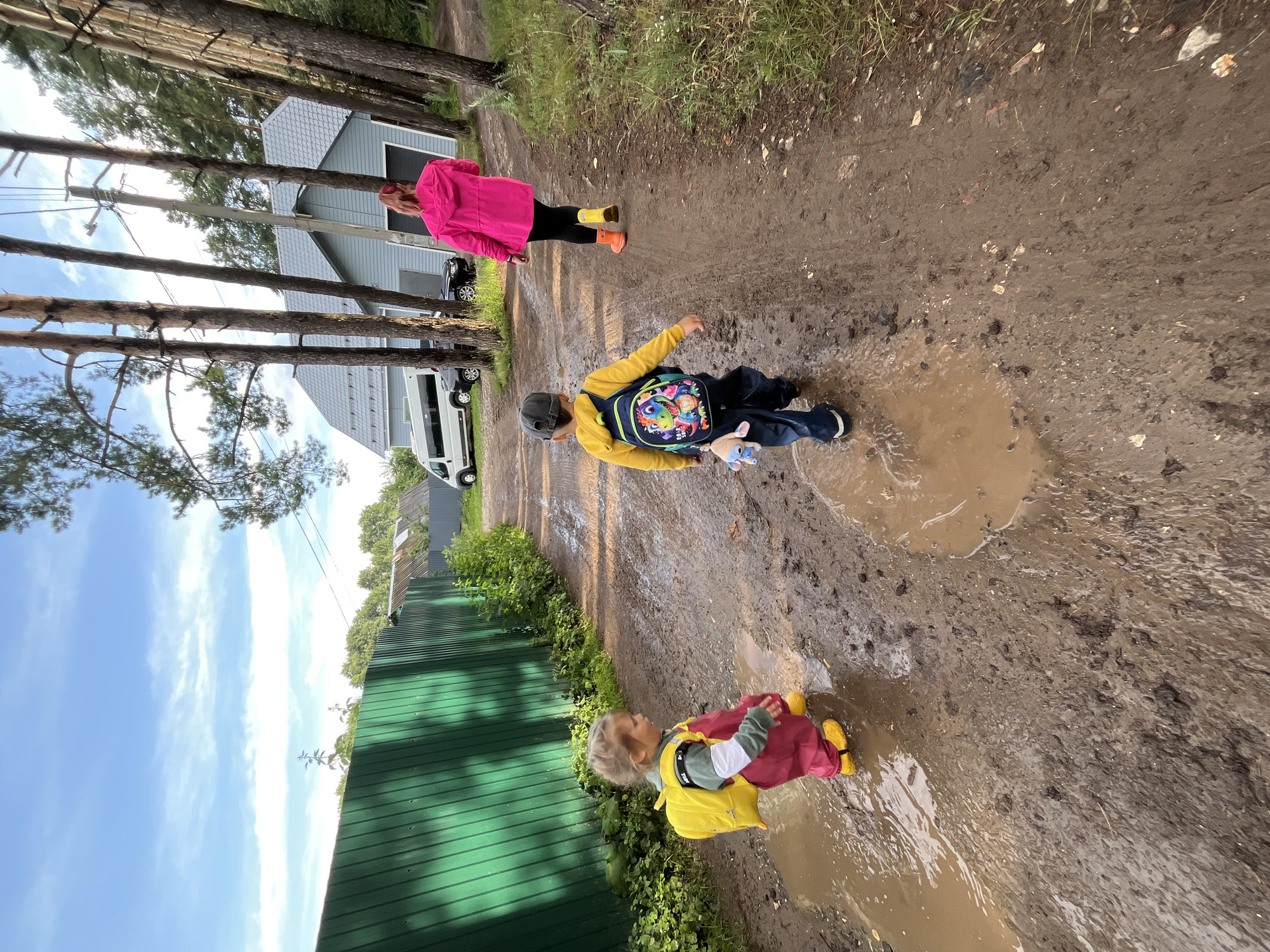 A young child wearing a yellow backpack and red overalls with yellow boots walks through a muddy puddle on a dirt road. Behind the child, another child in a yellow hoodie and blue jeans with a colorful backpack and a stuffed animal attached walks in the same direction. Further back, a person wearing a bright pink jacket and orange and yellow boots walks ahead, away from the camera. The scene is set in a rural area with a green fence on the left, trees lining the right side of the road, and a house with a gray roof in the background. A white van and a black car are parked near the house. The sky is partly cloudy. This scene is consistent with the outskirts of Chaikovsky, Russia, where such dirt roads and wooded areas are common.