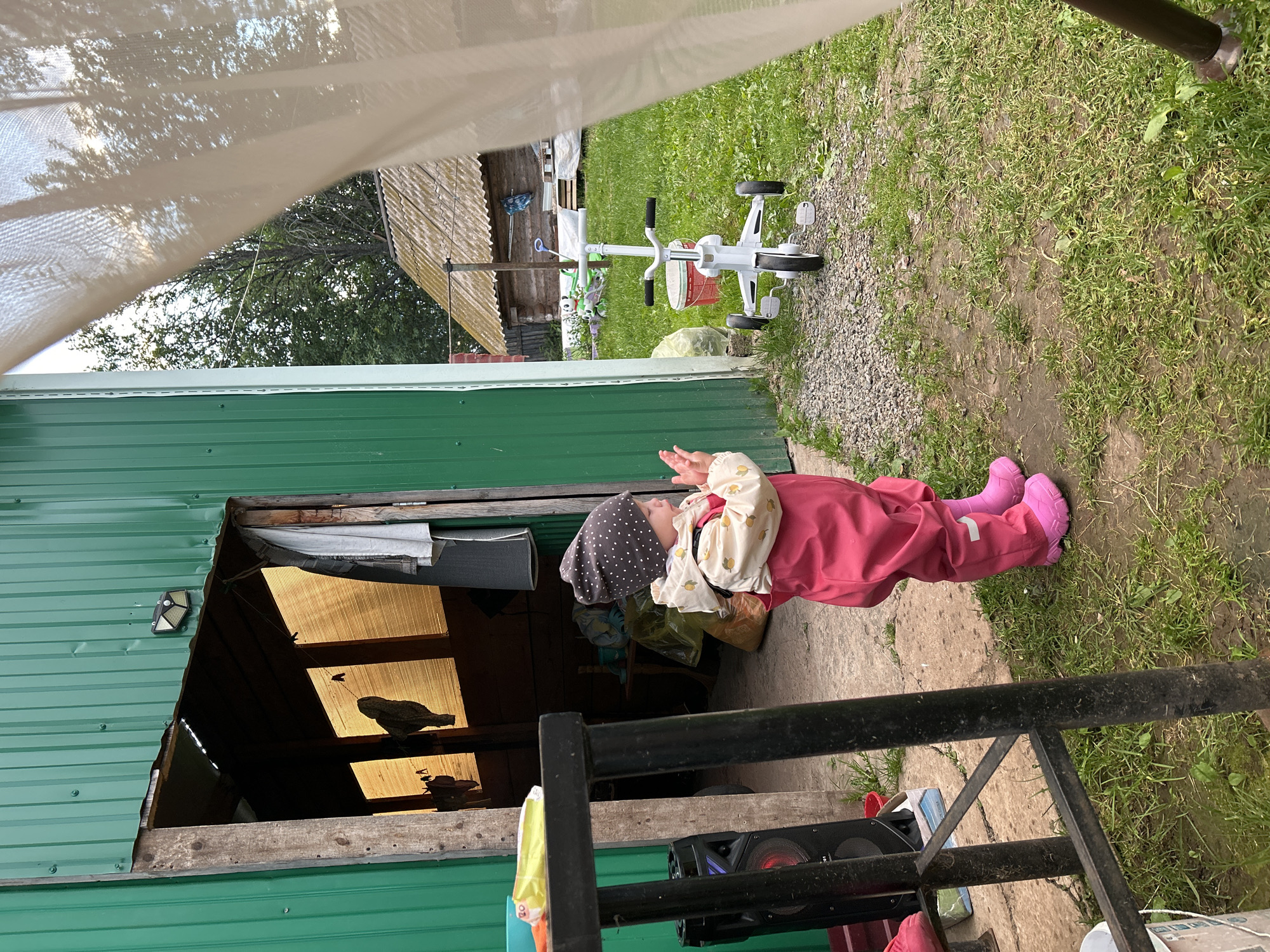 An outdoor, eye-level, medium shot captures a young child standing on a gravelly patch of ground in front of a green corrugated metal structure. The child, wearing a grey dotted hat, a patterned jacket, and pink overalls with matching pink boots, faces away from the camera and appears to be clapping their hands. A white child's tricycle with a red bucket is visible to the right of the child, on a grassy area. In the background, beyond the tricycle, is a wooden structure with a corrugated roof, and further beyond that, trees and a cloudy sky. To the left of the child, a black metal structure supports a black speaker with red accents. The ground is a mixture of gravel and grass, with some areas appearing damp. The overall scene suggests a rural or semi-rural outdoor setting. The location is identified as Azinsky, Russia.