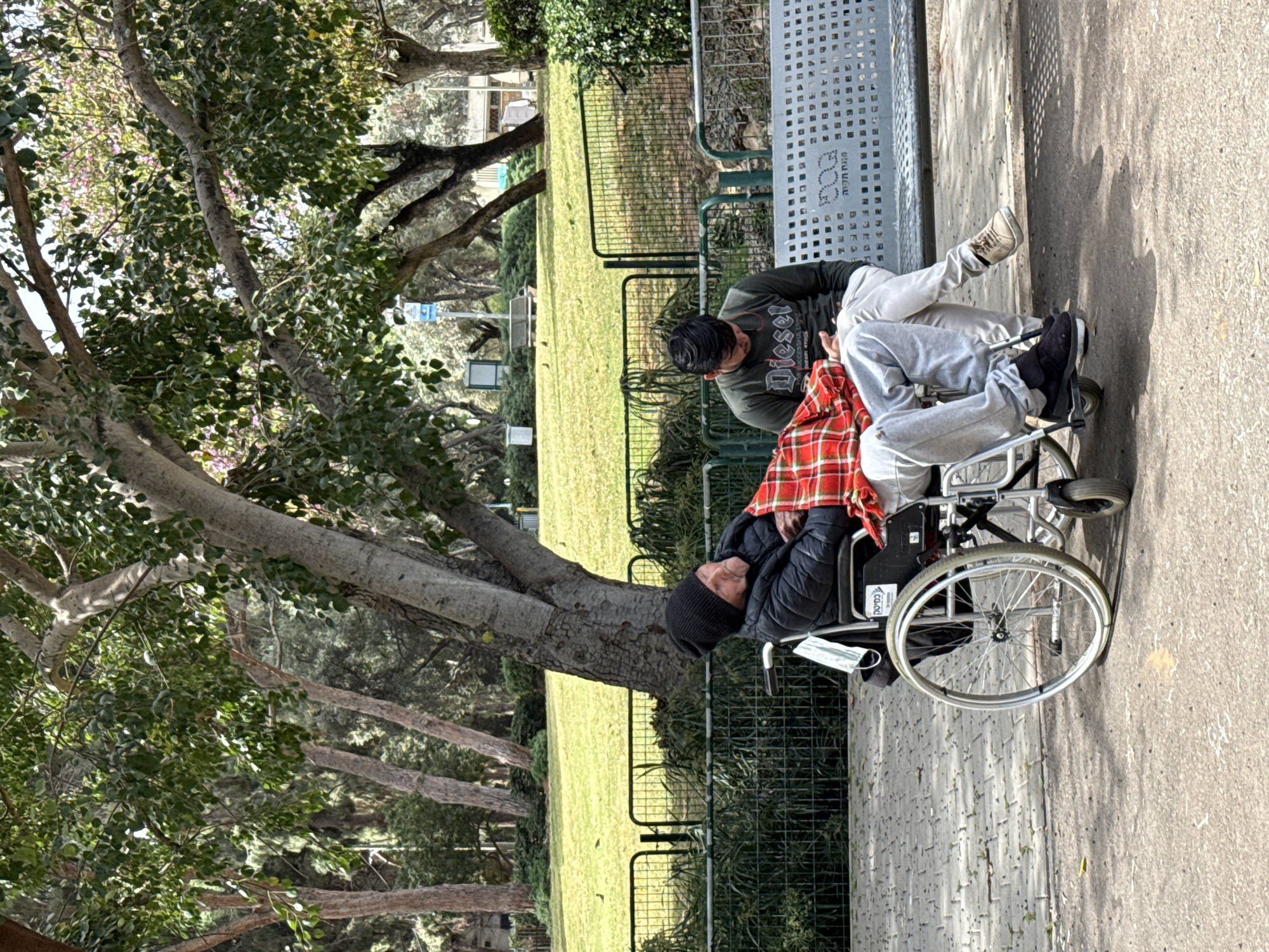 The image shows a person in a wheelchair, covered with a red and black plaid blanket, next to a younger person. The person in the wheelchair is wearing a black beanie and dark clothing. The younger person is wearing a dark green shirt with the word "Diesel" printed on it and light-colored sweatpants. They are positioned next to a metal bench in a park. The background includes a grassy area, trees, and other foliage. The scene appears to be in a public park. The image was likely taken in Petah Tikva, Israel, based on the text visible on the bench.