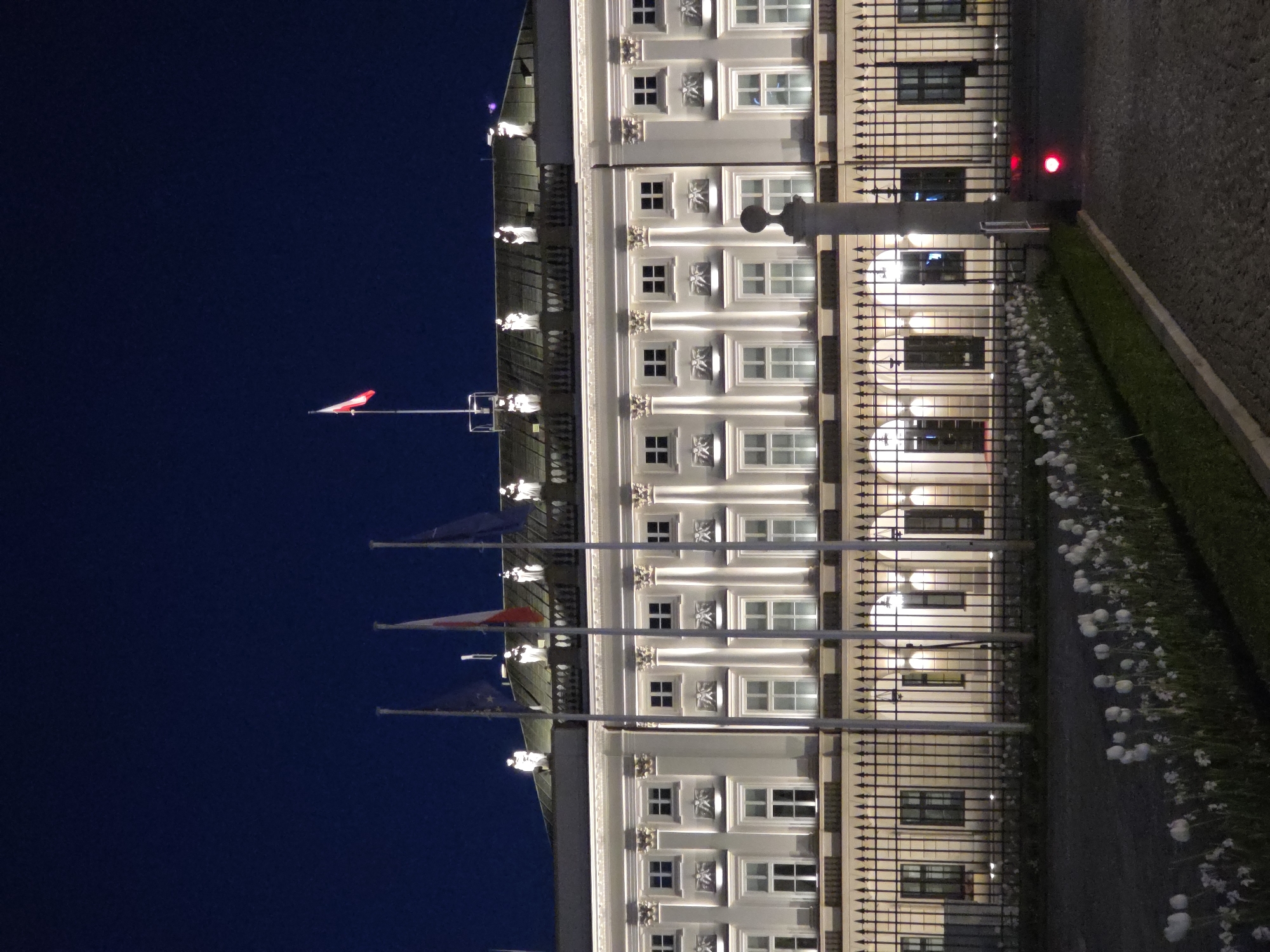 The image depicts the Presidential Palace in Warsaw, Poland at night. The building's facade is illuminated, showing windows and decorative architectural elements. Flags on poles are visible against a dark blue sky. A wrought-iron fence runs along the front of the palace, with flowers and greenery alongside a paved area. A red light is visible near the fence. No people are visible in the frame.