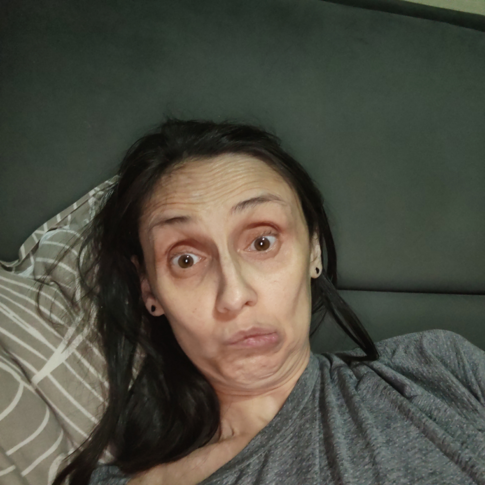 A woman makes a goofy face as she takes a selfie from a bed. Her head is turned slightly to the left, and her eyes are wide open and looking at the camera. Her mouth is puckered, and her chin is pulled up, creating a double chin. She has dark hair that is falling around her face, and she is wearing a gray t-shirt. The background is a dark green wall, and a pillow with a striped pattern is visible behind her head.