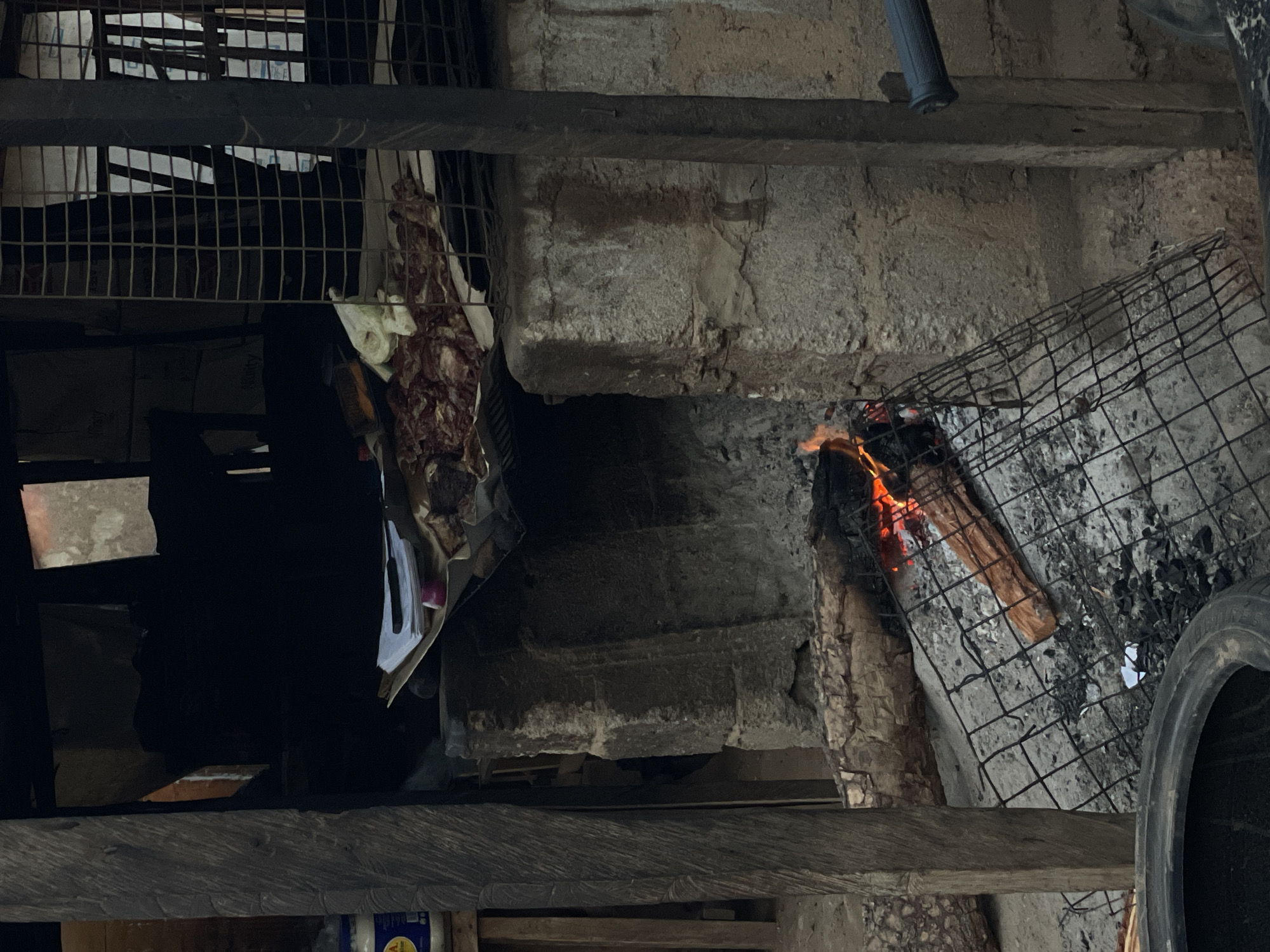 A scene of an improvised cooking area in Akwanga, Nigeria. It includes a rough, stone-built structure, likely serving as a makeshift hearth or oven. There's a fire burning with visible flames and smoldering embers inside a wire mesh grill. Pieces of wood fuel the fire. There's another, larger, rusty wire cage structure located above the hearth. A bundle of meat and scraps of paper or plastic hang from the cage, indicating cooking activities. The environment is utilitarian, with gray concrete, wood and grime.