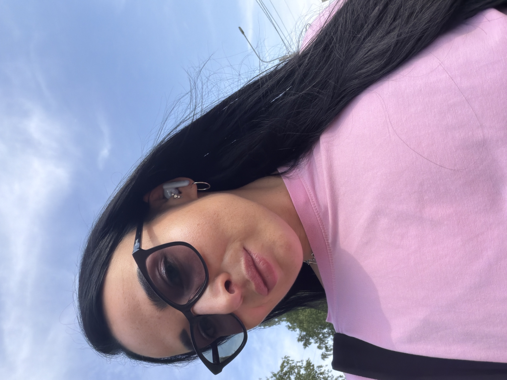 A woman with dark hair and sunglasses poses close to the camera. She wears a pink top. There is a blue sky in the background. Based on the style of the street lighting and overall environment, this is likely in Saint Petersburg, Russia.