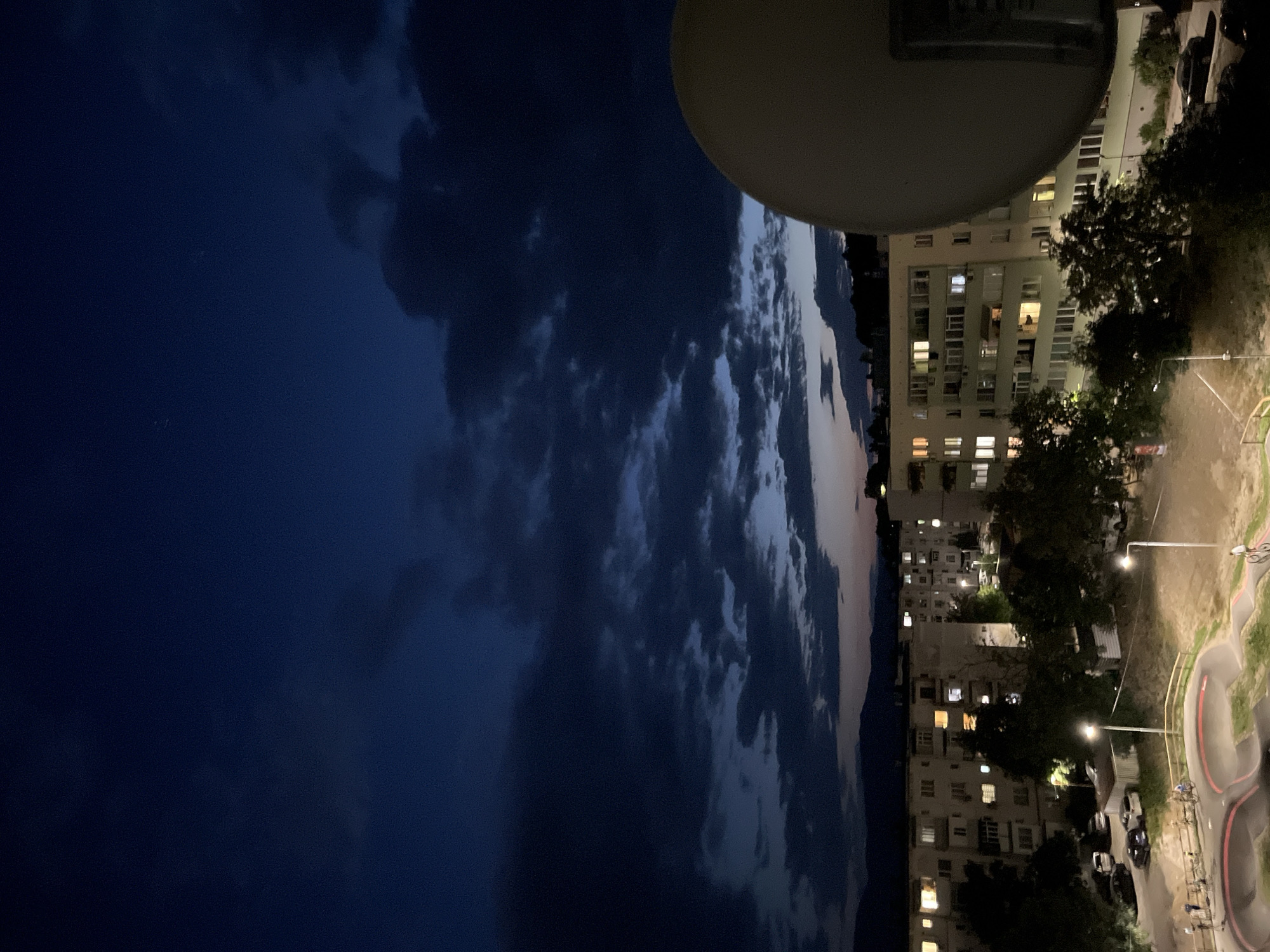 This is a nighttime photograph taken from an elevated perspective, likely a balcony or rooftop, overlooking a residential area in Sopot, Bulgaria. The sky is a deep, dark blue with dramatic, heavy clouds that appear to be illuminated from below by the city lights. In the foreground, a large, pale satellite dish occupies the right side of the frame. Below the dish, apartment buildings with illuminated windows are visible, suggesting it is evening or early night. Trees are scattered around the buildings. In the mid-ground, a paved area with a circular track, possibly a pump track for bicycles or skateboards, is illuminated by streetlights. Several people are visible on the track, some with bicycles, indicating recreational activity. Cars are parked in a lot near the buildings. In the background, a faint silhouette of mountains can be seen against the twilight sky. The overall scene depicts a segment of urban life in Sopot during the evening.