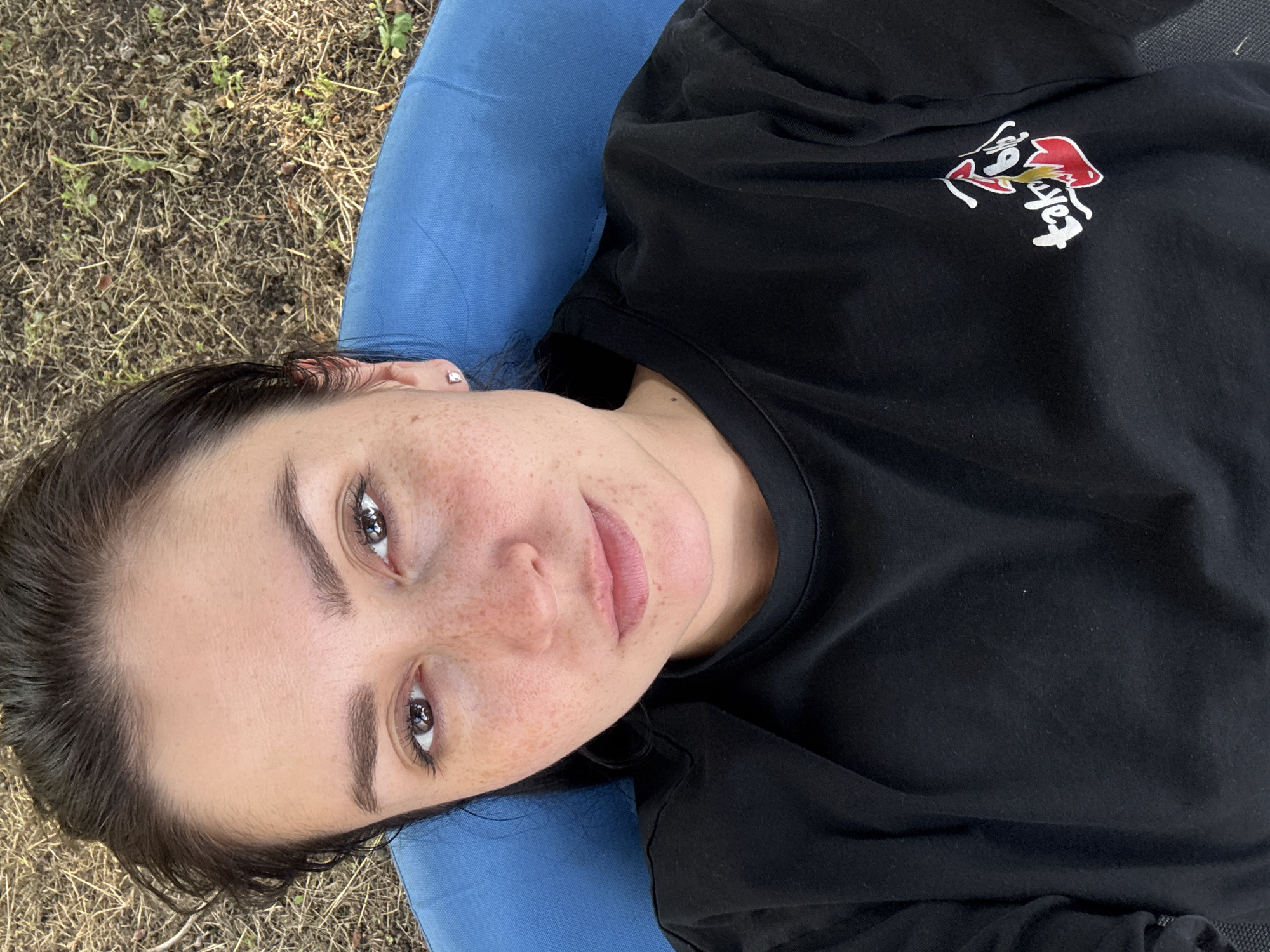 A close-up, overhead shot shows a woman lying down outdoors, looking up at the camera. She has dark hair, light skin with freckles, and is wearing a black t-shirt. She is partially reclined on a blue cushioned surface, which is set against a backdrop of dry grass and dirt. The image is sharp and well-lit, with the woman's face as the primary focus. The scene appears to be in a natural outdoor setting, possibly a park or backyard. There are no other people or animals visible in the frame. The overall impression is one of casual relaxation in an outdoor environment.