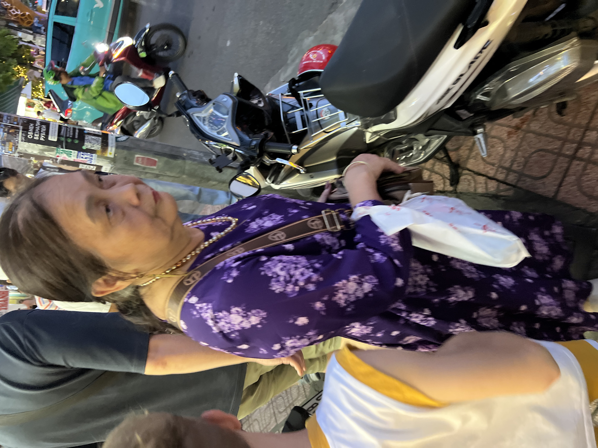 An outdoor, eye-level medium shot captures a woman in her 60s in a vibrant purple floral dress standing on a sidewalk in Nha Trang, Vietnam. She is positioned slightly to the left of center, her head turned away from the camera. Her hair is dark with streaks of gray pulled back into a ponytail. A gold necklace adorns her neck, and a brown crossbody bag with a patterned strap is slung over her shoulder. She carries a white plastic bag in her left hand. To the right of the woman, a silver and black scooter is parked, its seat and part of the front fairing visible. A red helmet rests on the rear of the scooter. In the background, a person in a green jacket rides another scooter, their face obscured by a green helmet. Behind them, a teal-colored vehicle with the branding "GRAB" is visible. The scene is set against a backdrop of a street with cobblestone pavement, a lamppost with posters, and building facades with lights. The overall impression is a snapshot of everyday life in a bustling Vietnamese city.