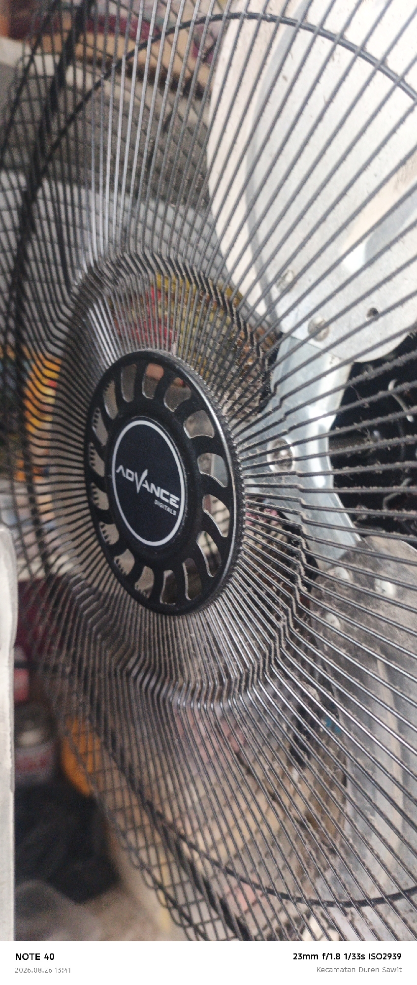 The image is a close-up, slightly angled shot of a fan's protective grille and central motor housing. The fan grille is made of thin black wires, forming a dense mesh. The central part of the fan, where the "ADVANCE DIGITALS" logo is visible in white lettering on a black circle, is also a focal point. The background is blurred but suggests an indoor setting with some colorful objects and possibly other appliances. The lighting appears to be artificial, casting shadows through the fan grille. The overall impression is one of a domestic appliance in a slightly dusty or lived-in environment. The image includes text overlays indicating "NOTE 40", "2026.08.26 13:41", "23mm f/1.8 1/33s ISO2939", and "Kecamatan Duren Sawit". While the text mentions "Kecamatan Duren Sawit", which is a subdistrict in East Jakarta, Indonesia, the image itself does not provide visual cues to confirm this specific location.