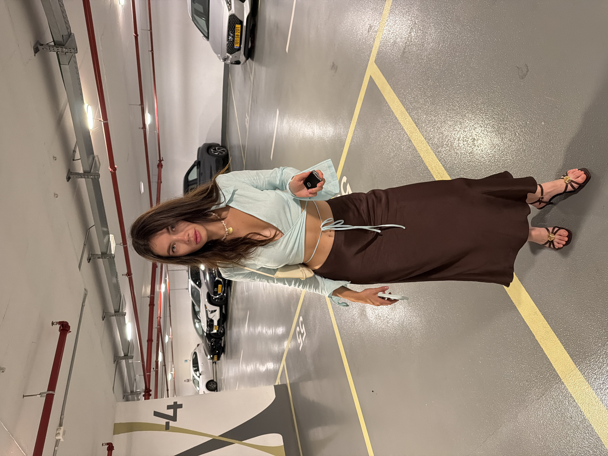 A woman is standing in a dimly lit parking garage, possibly in Tel-Aviv, Israel. The floor is concrete with yellow parking lines. To her left, a large white pillar is marked with a black number "4". Several cars are parked in the background, including a silver Hyundai on the right side of the frame with an Israeli license plate. The woman is wearing a light blue cropped wrap top with bell sleeves, a brown satin maxi skirt, and dark strappy sandals with gold embellishments. She has a small beige purse slung over her shoulder and is holding a car key in her right hand and a white object, possibly a phone, in her left. She is wearing a pearl necklace with a shell pendant.