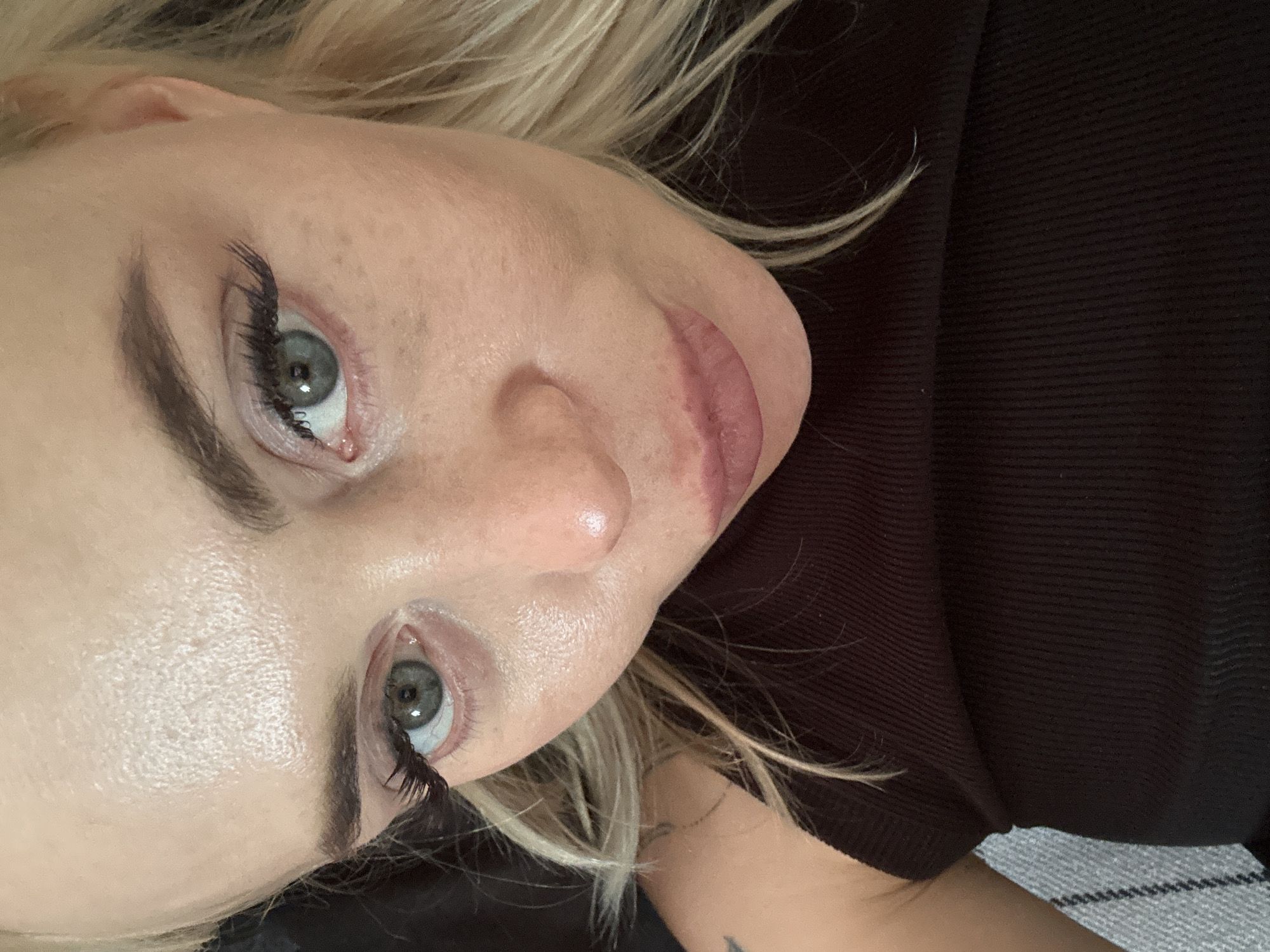 A close-up, eye-level portrait depicts an adult female with blonde hair and light-colored eyes, looking directly into the camera. Her skin exhibits a slight sheen, particularly on the forehead, nose, and cheeks, with visible pores and minor freckling around the eyes. Her eyebrows are dark and well-defined. Both eyes are accentuated with dark eyeliner and artificial eyelashes; her irises are a blue-green color. Her lips are partially open, coated with a muted pink lipstick. She is wearing a dark, possibly black or very dark brown, vertically ribbed sleeveless garment. A portion of her left shoulder and upper arm is visible, revealing a dark mark or small tattoo. In the background, to the left of her arm, a textured grey and white fabric, possibly upholstery, is partially visible. The remainder of the background is dark and indistinct, suggesting an indoor setting. The location is the city of Unknown, Unknown.