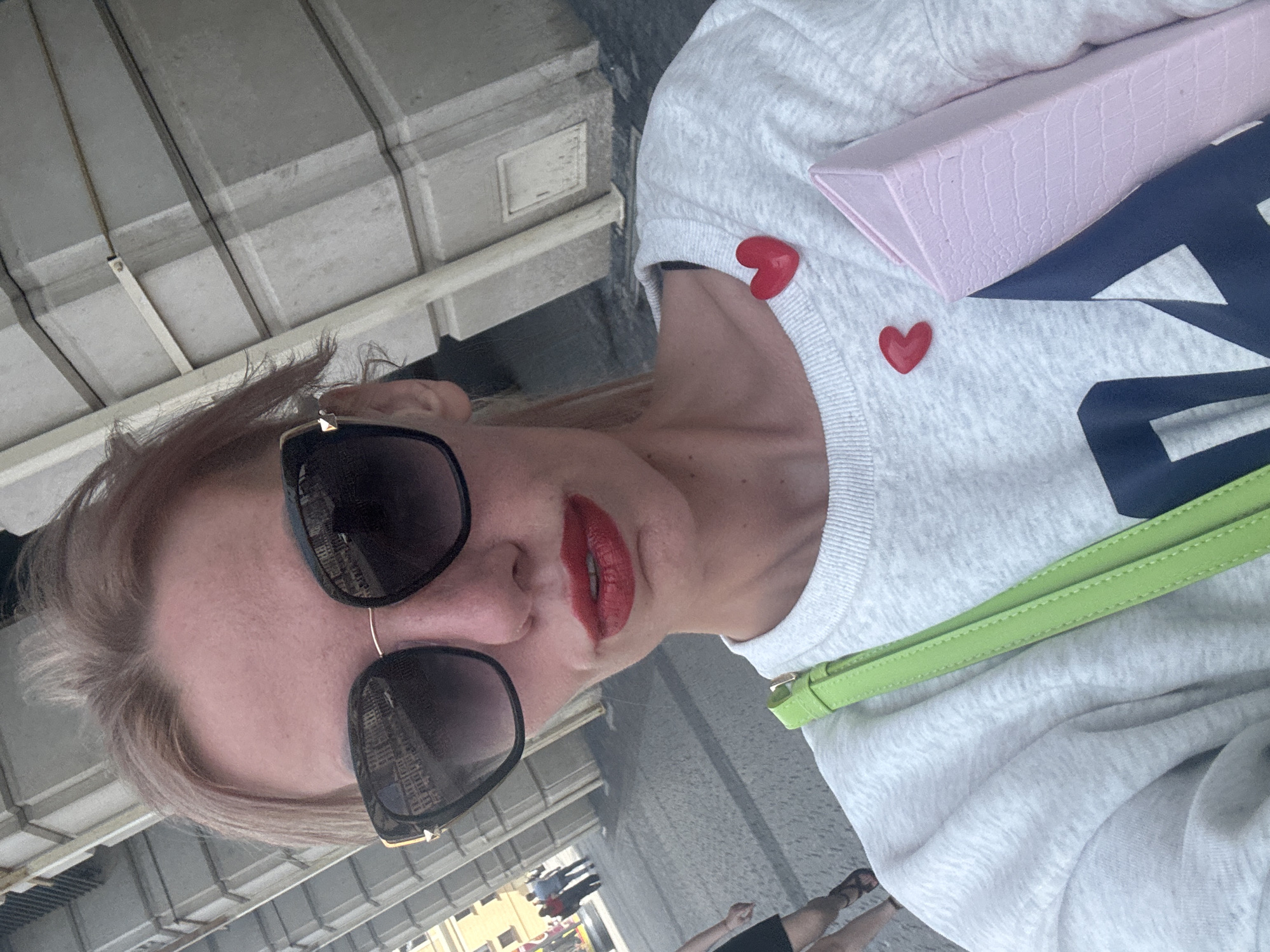 The image is a close-up selfie of a woman with blonde hair, wearing sunglasses and red lipstick. She has two red heart-shaped pins on her light gray sweatshirt, and a lime green strap from a bag is visible. In the background, there are large stone columns, suggesting an architectural setting. The ground appears to be paved with cobblestones, typical of city streets. Several figures are visible in the distance, walking along the street. Based on the architecture with its classical columns and the overall urban environment, this scene is likely located in the historical center of Saint Petersburg, Russia, possibly near one of its grand avenues or squares, such as Nevsky Prospekt or Palace Square. The sunlight indicates it is daytime.
